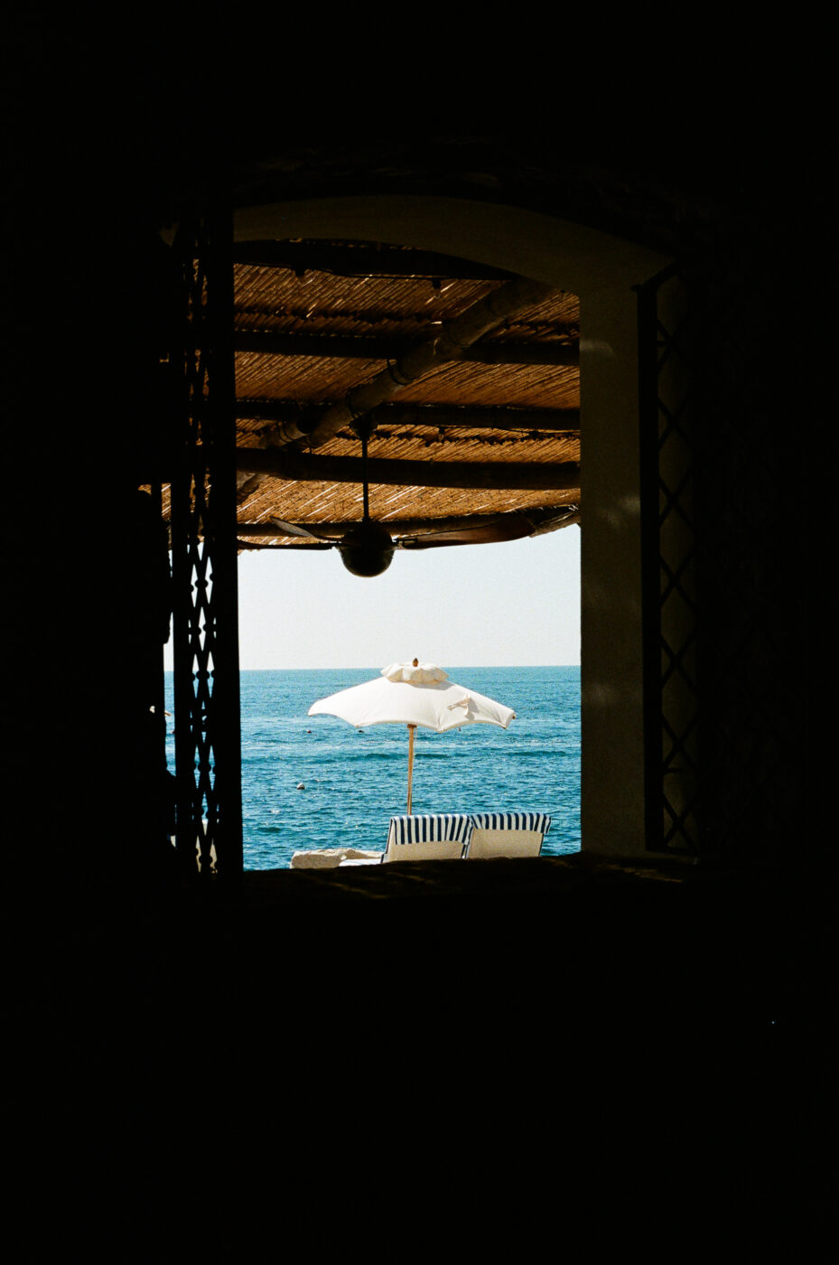 Una vista attraverso la cornice di una finestra scura rivela un patio illuminato dal sole con un ombrellone bianco, sedie a sdraio e l'oceano blu sullo sfondo sotto un cielo limpido. (Nota: poiché non è stato fornito il titolo dell'immagine, il testo alt inizia direttamente con la descrizione, secondo le vostre linee guida di formattazione).