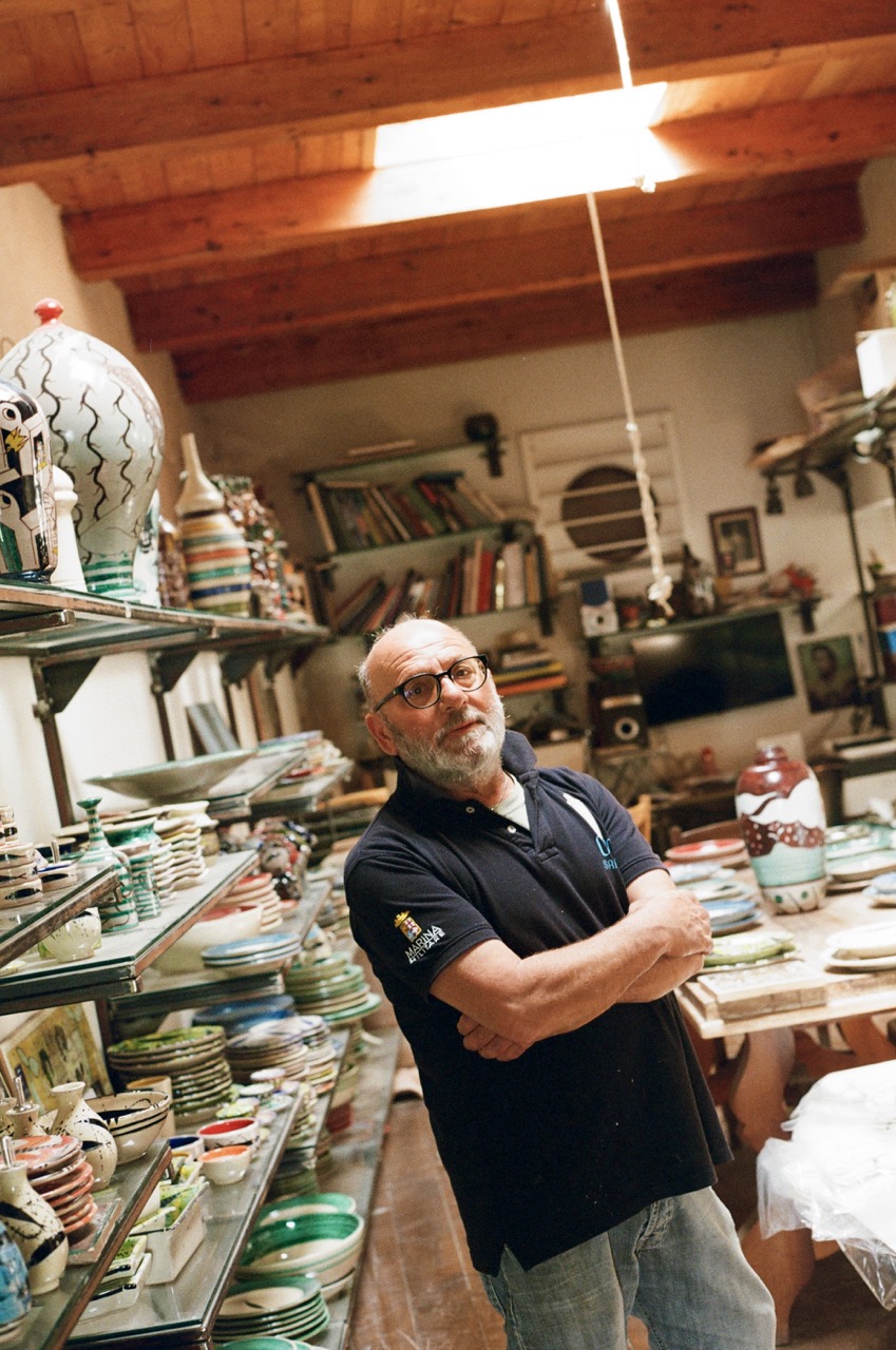 Non è stato fornito alcun titolo dell'immagine, quindi il testo alt appropriato sarebbe: Un uomo con barba bianca e occhiali sta a braccia conserte in uno studio di ceramica pieno di ceramiche colorate e scaffali di piatti, ciotole e vasi. La luce del sole entra da una finestra in alto.