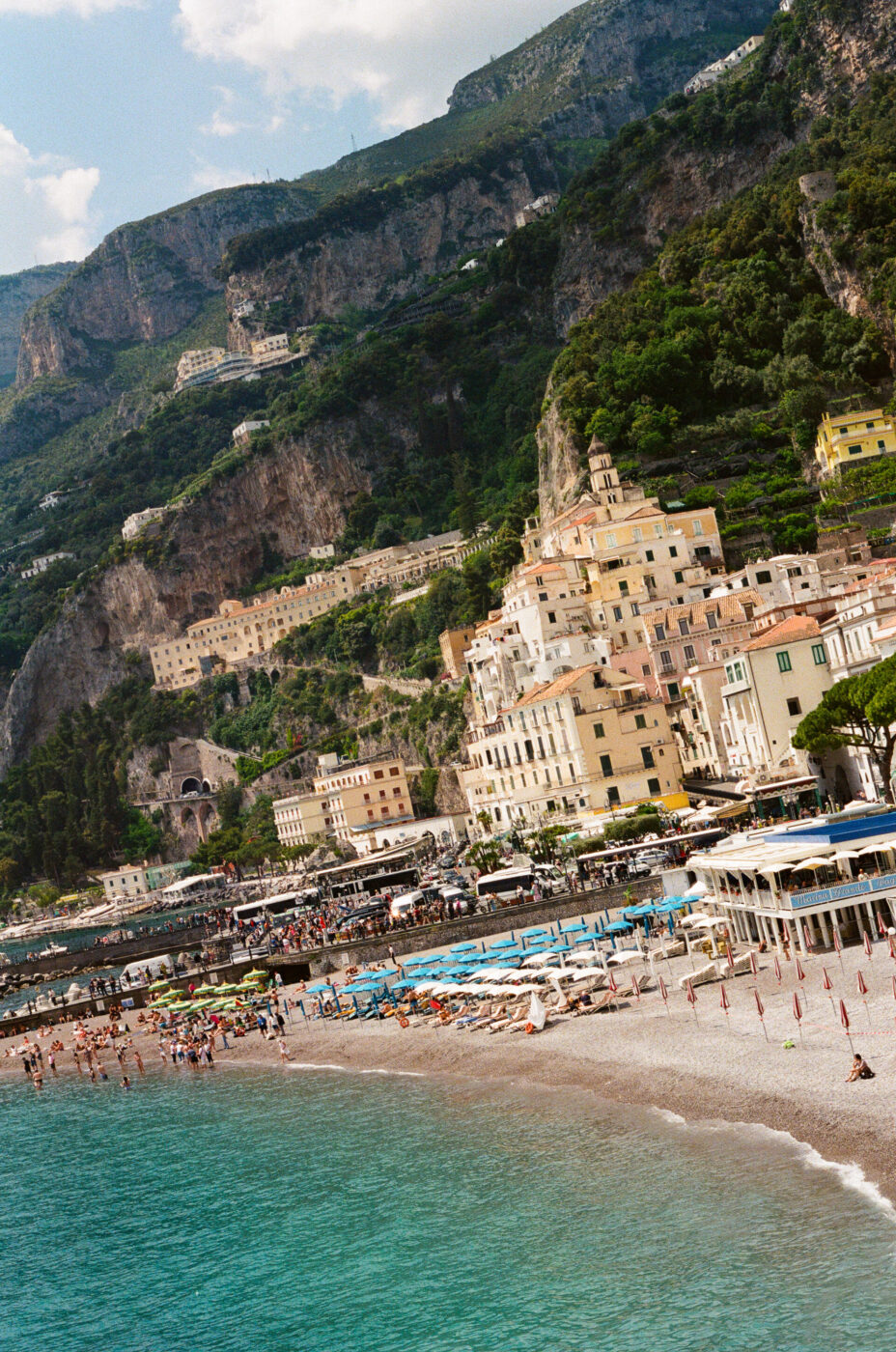 Città costiera panoramica - Una città costiera panoramica con edifici colorati lungo il pendio di una collina, una spiaggia di ciottoli affollata con ombrelloni e bagnanti e acqua turchese, incastonata in scogliere verdeggianti.