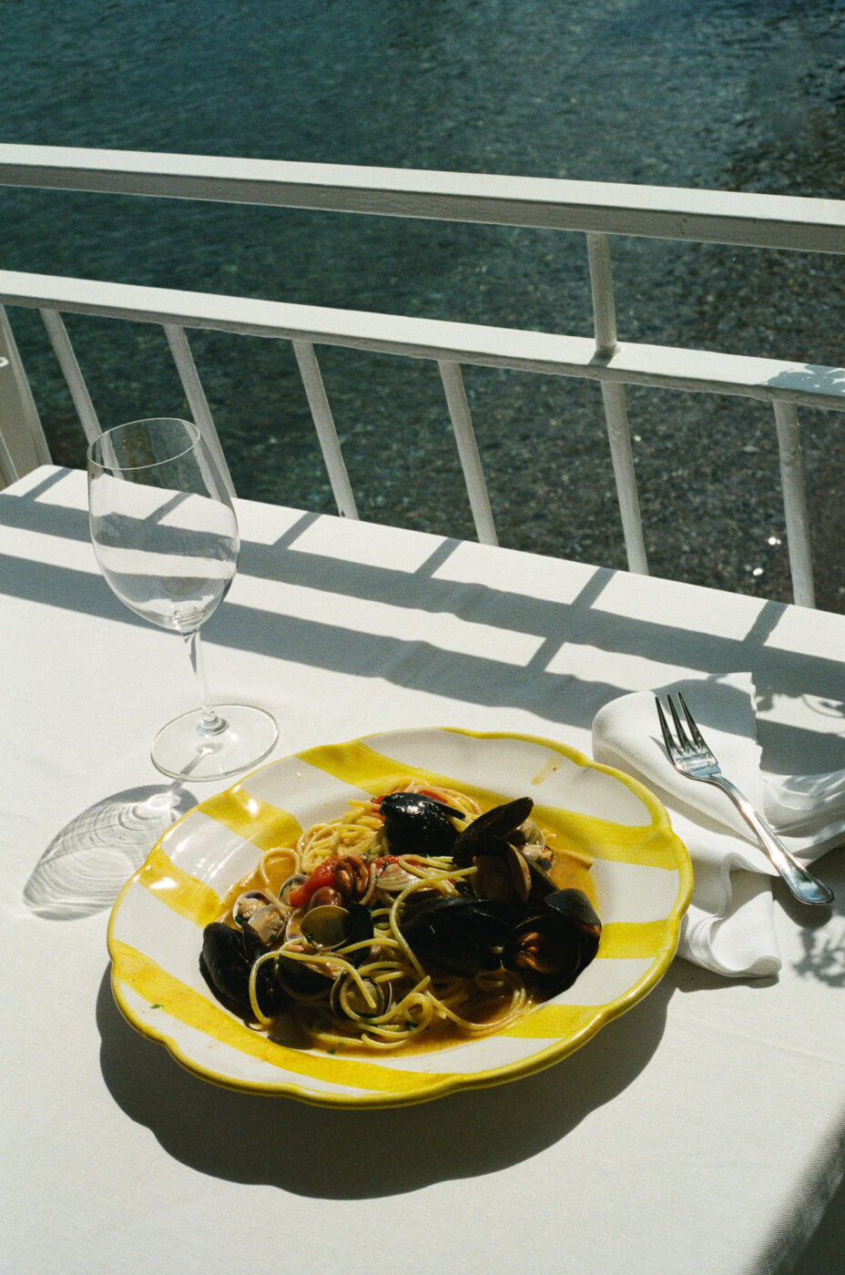 Titolo immagine non fornito - Un piatto di pasta ai frutti di mare si trova su una tovaglia bianca accanto a una forchetta, un tovagliolo e un bicchiere di vino vuoto, con la luce del sole che proietta ombre e l'acqua visibile attraverso una ringhiera bianca sullo sfondo.