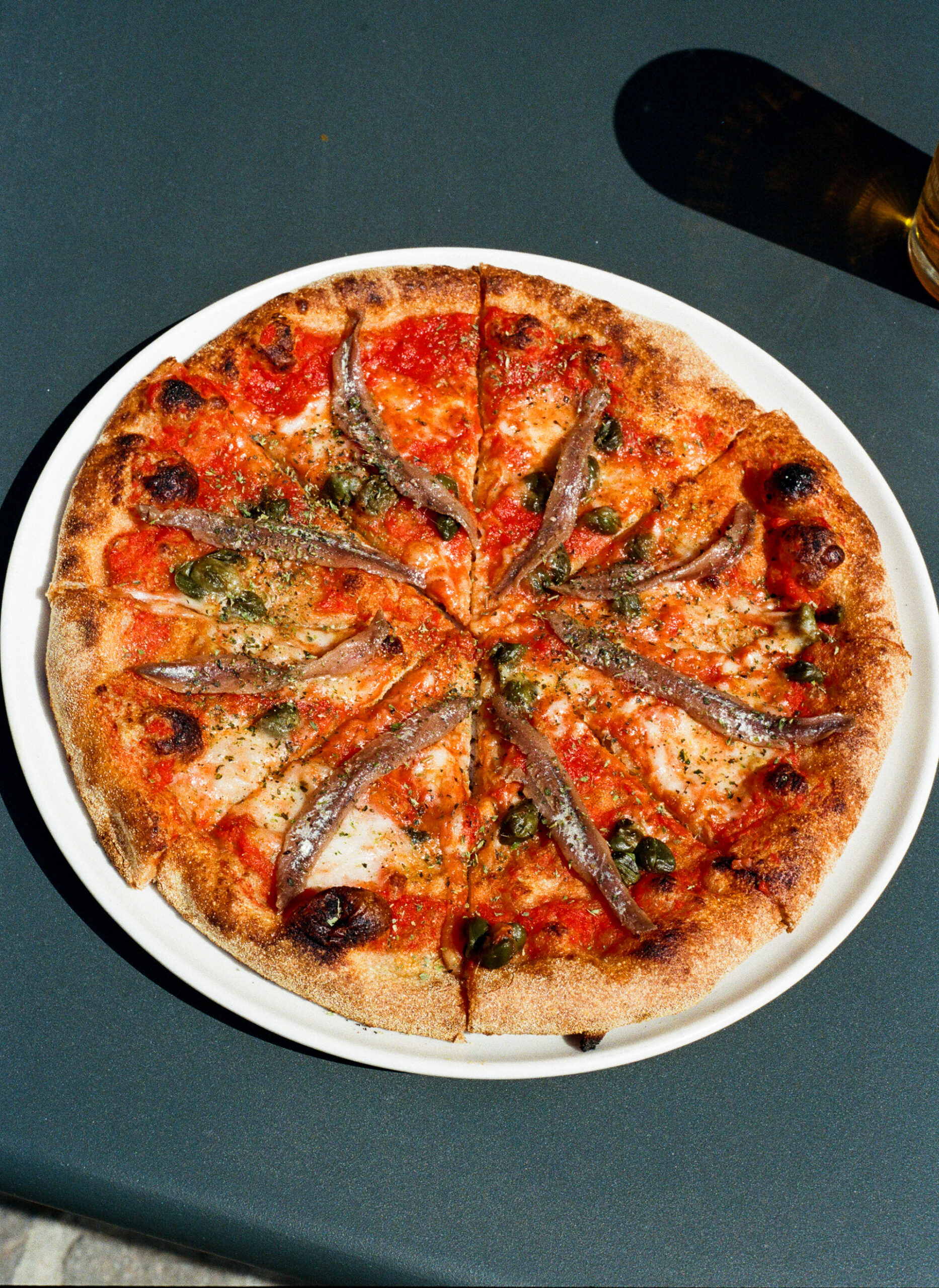 Pizza - A round pizza topped with tomato sauce, melted cheese, anchovies, and herbs is sliced into eight pieces on a white plate, sitting on a dark table.