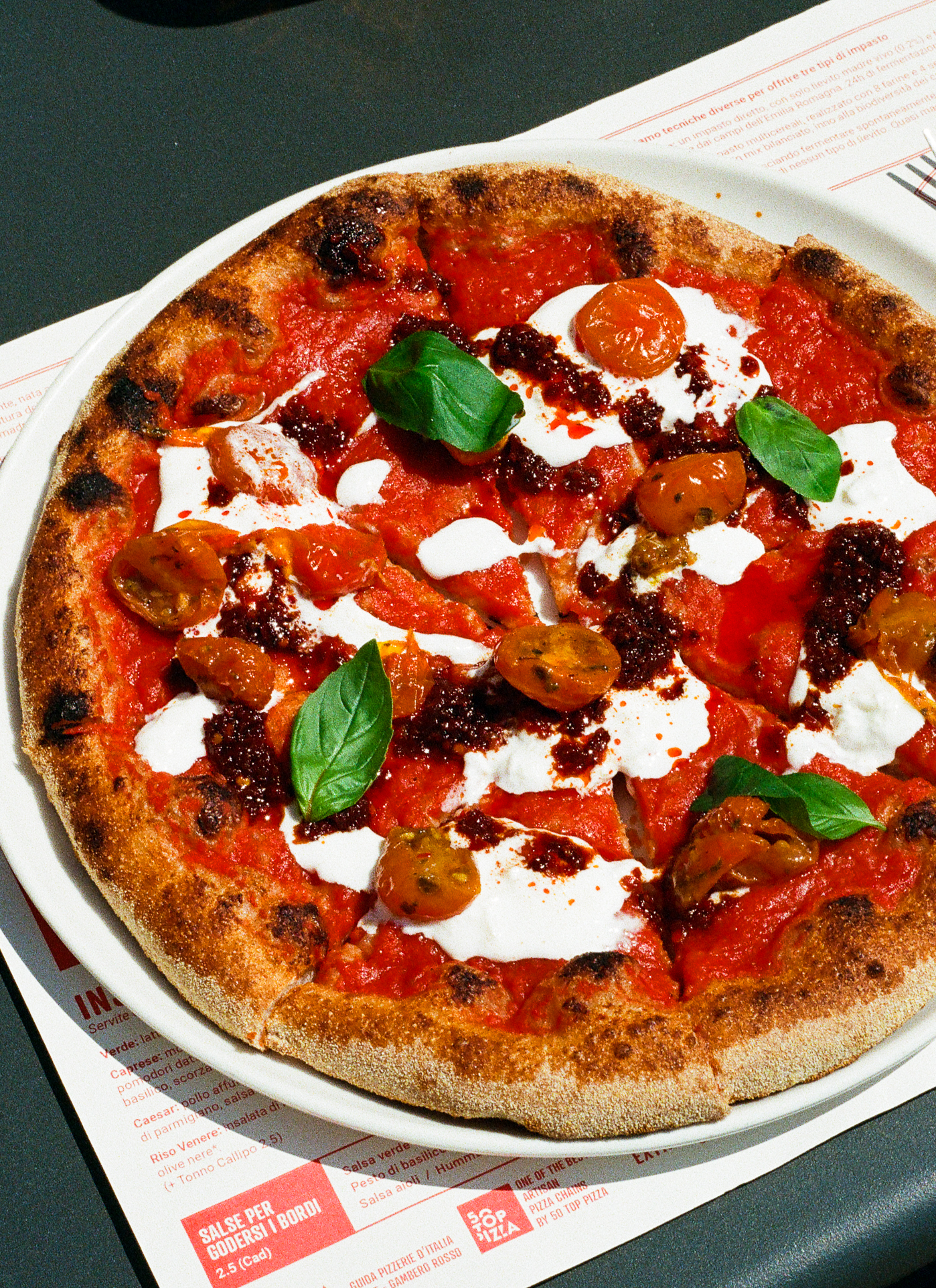 A rustic pizza topped with tomato sauce, dollops of cheese, halved cherry tomatoes, spicy red oil, and fresh basil leaves, served on a white plate atop a menu.