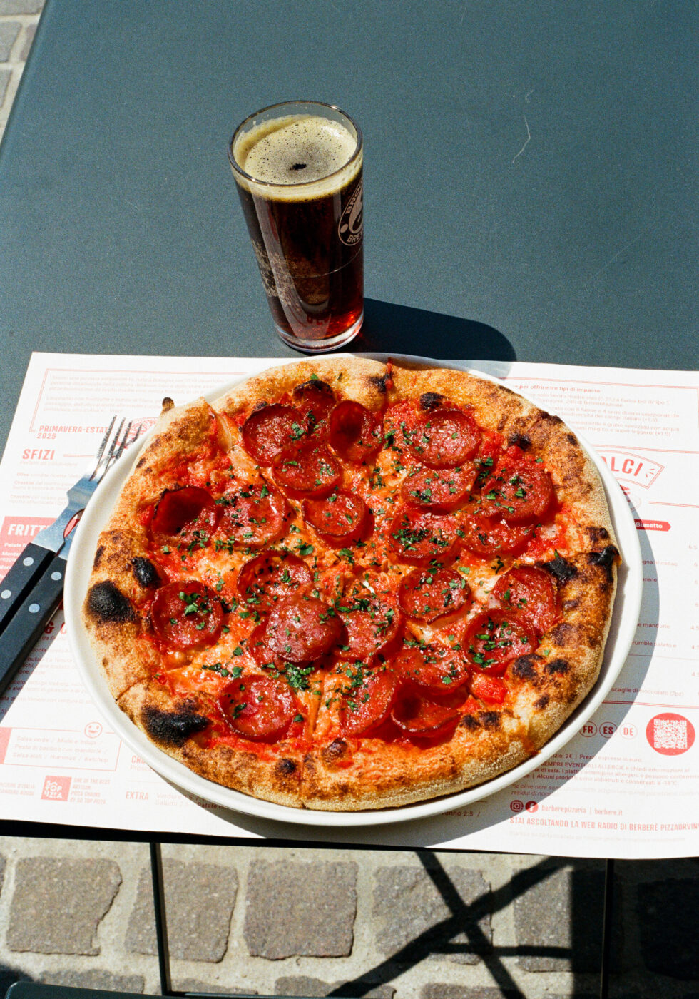A pepperoni pizza on a white plate sits on a table next to a glass of dark beer, with utensils and a menu visible underneath, outdoors on a sunny day. If there is no specified image title present, you should just use: A pepperoni pizza on a white plate sits on a table next to a glass of dark beer, with utensils and a menu visible underneath, outdoors on a sunny day.