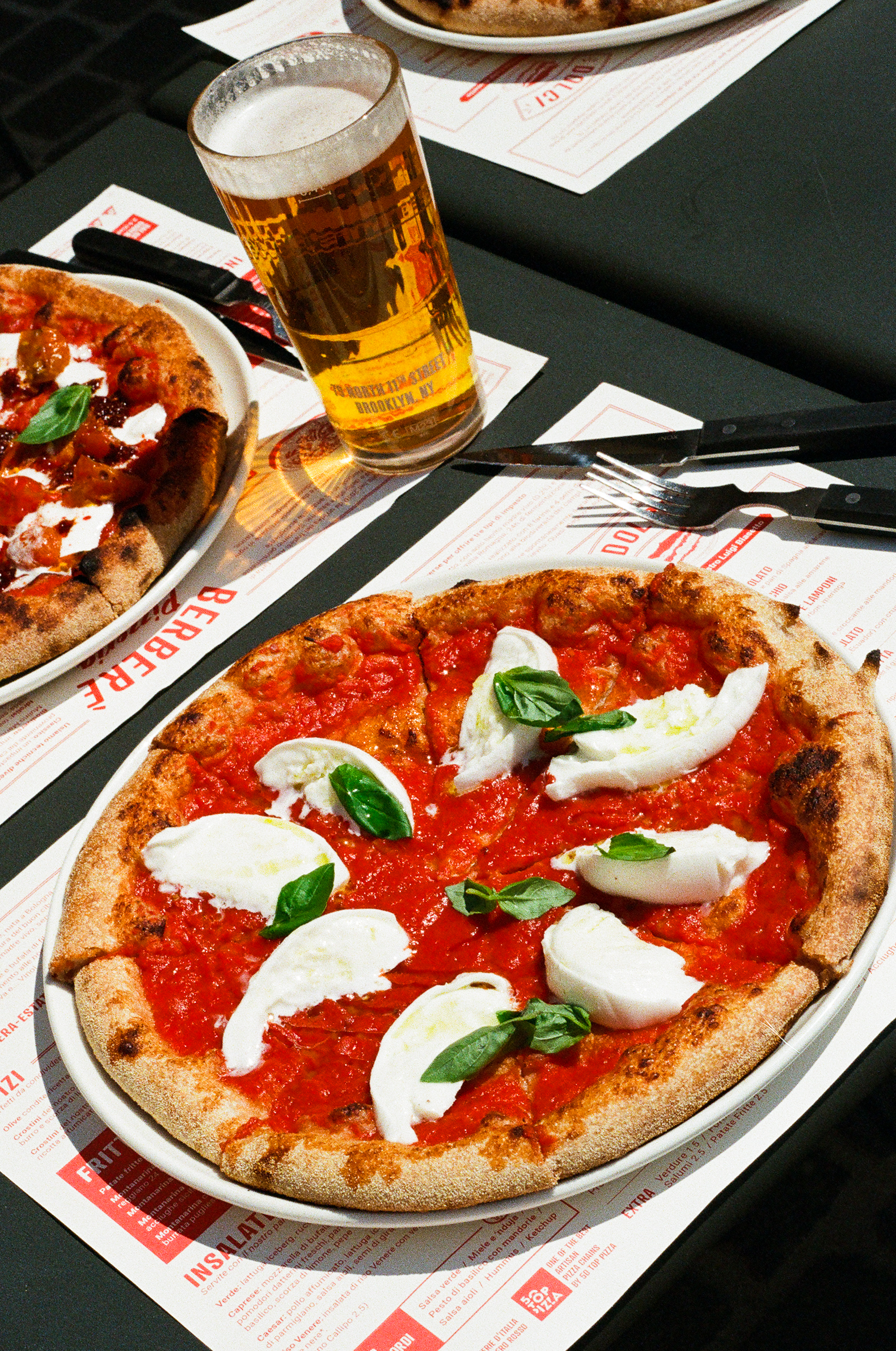 Margherita Pizza and Beer - A plate of pizza topped with tomato sauce, mozzarella slices, and fresh basil sits on a table next to a glass of beer and utensils, with another pizza visible in the background.
