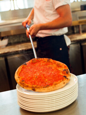 A chef in a white shirt uses a metal pizza peel to place a freshly baked pizza topped with tomato sauce on a stack of white plates in a kitchen. *(Note: You did not provide an image title, so the alt text starts directly with the description. If theres an image title, please specify it and I will format accordingly.)*.