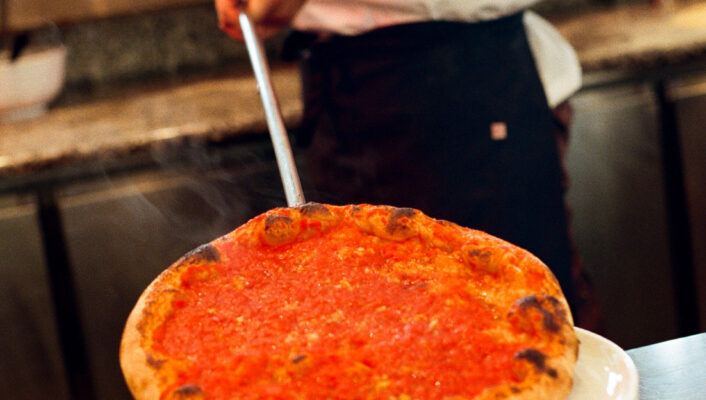 A chef in a white shirt uses a metal pizza peel to place a freshly baked pizza topped with tomato sauce on a stack of white plates in a kitchen. *(Note: You did not provide an image title, so the alt text starts directly with the description. If theres an image title, please specify it and I will format accordingly.)*.