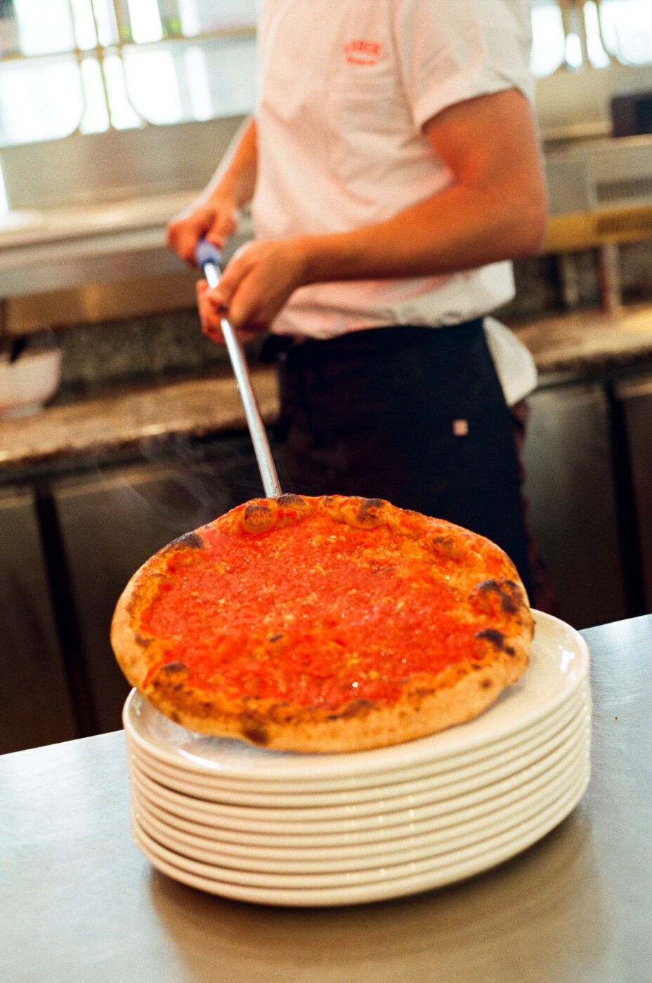 A chef in a white shirt uses a metal pizza peel to place a freshly baked pizza topped with tomato sauce on a stack of white plates in a kitchen. *(Note: You did not provide an image title, so the alt text starts directly with the description. If theres an image title, please specify it and I will format accordingly.)*.