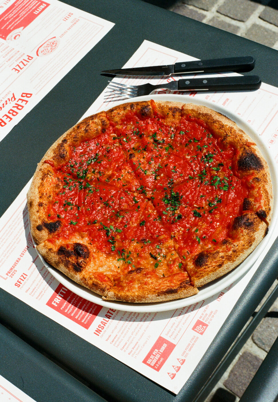 Pizza su piatto bianco: un'intera pizza condita con salsa di pomodoro ed erbe aromatiche viene servita su un piatto bianco, appoggiato su un menu sopra un tavolo scuro, con un coltello e una forchetta a portata di mano.