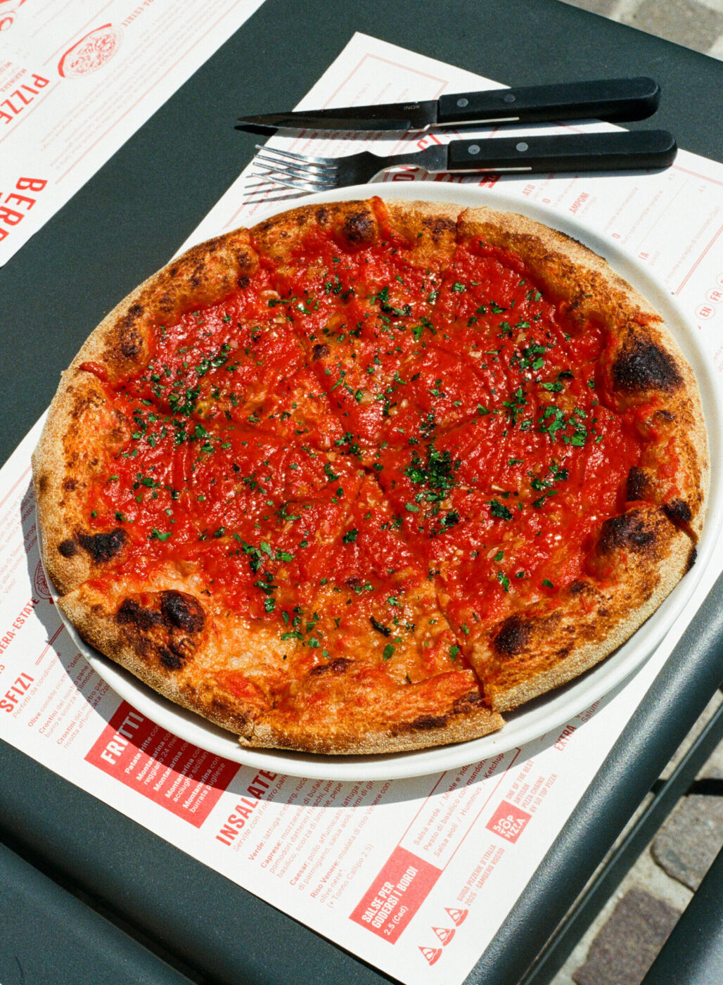 A round pizza topped with tomato sauce and herbs is served on a white plate, placed on a table with menus, alongside a fork and knife. The crust is golden and slightly charred.