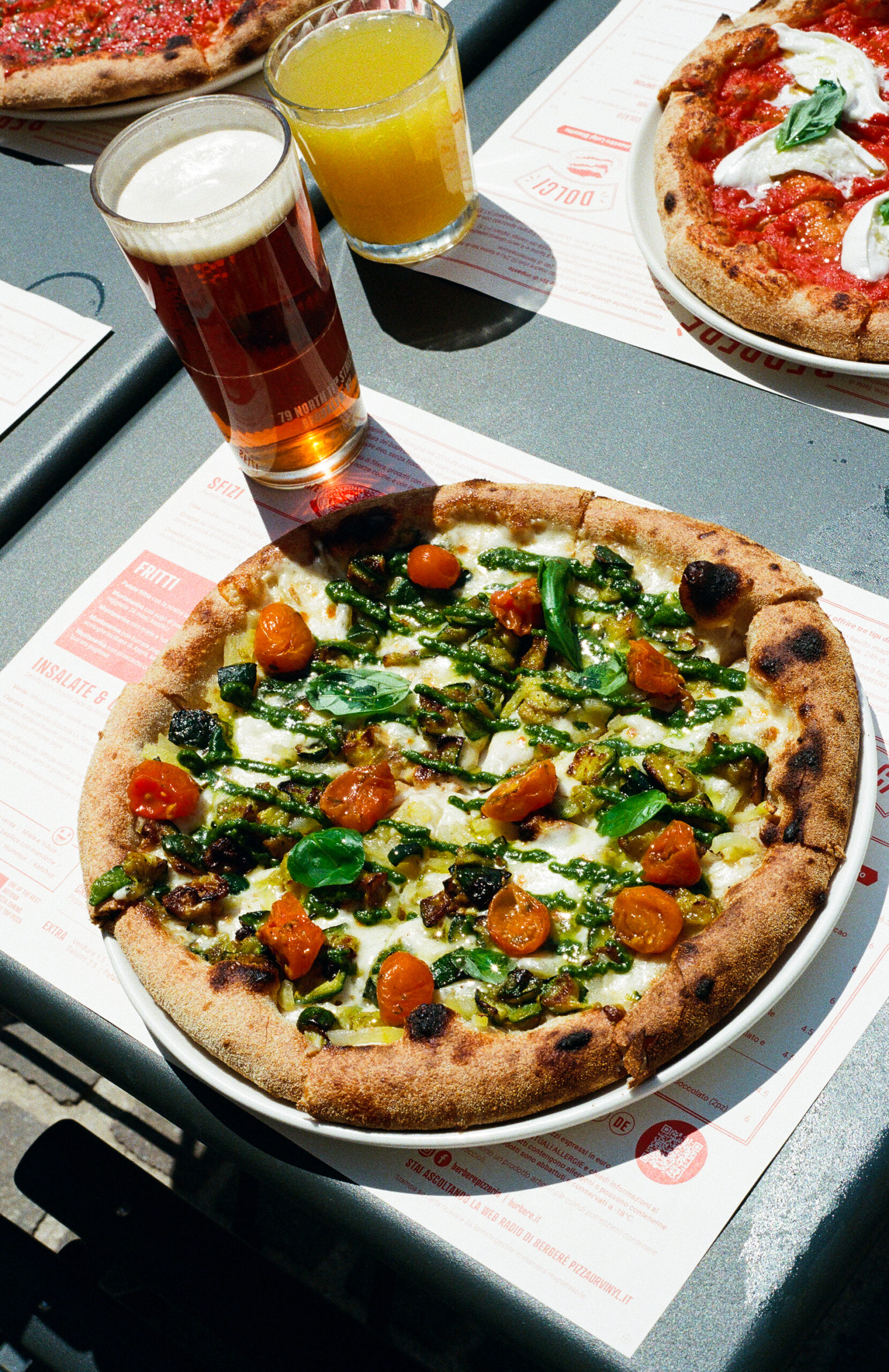 A wood-fired pizza topped with cherry tomatoes, greens, and cheese sits on a plate next to a glass of beer and a glass of orange juice on an outdoor table with other pizzas in the background. If there’s no image title, just use: A wood-fired pizza topped with cherry tomatoes, greens, and cheese sits on a plate next to a glass of beer and a glass of orange juice on an outdoor table with other pizzas in the background.