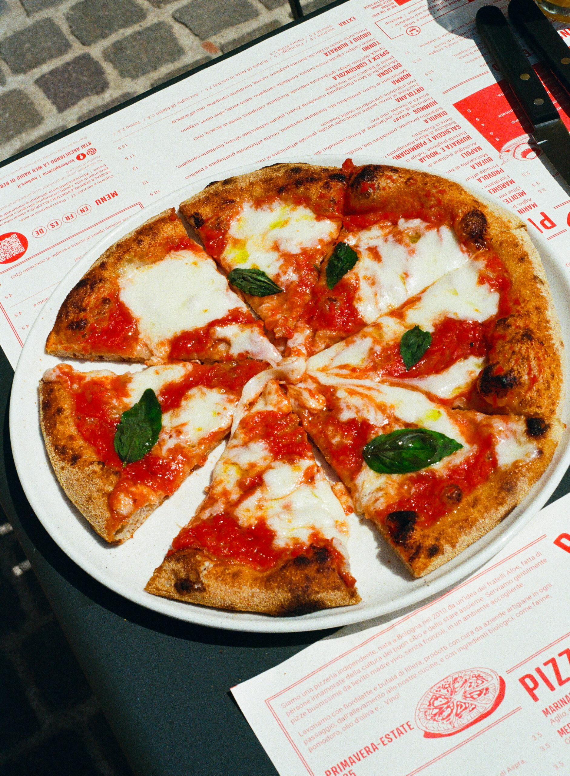Margherita Pizza - A Margherita pizza with melted mozzarella, tomato sauce, and fresh basil leaves is sliced into eight pieces and served on a white plate atop a table with red and white menus.
