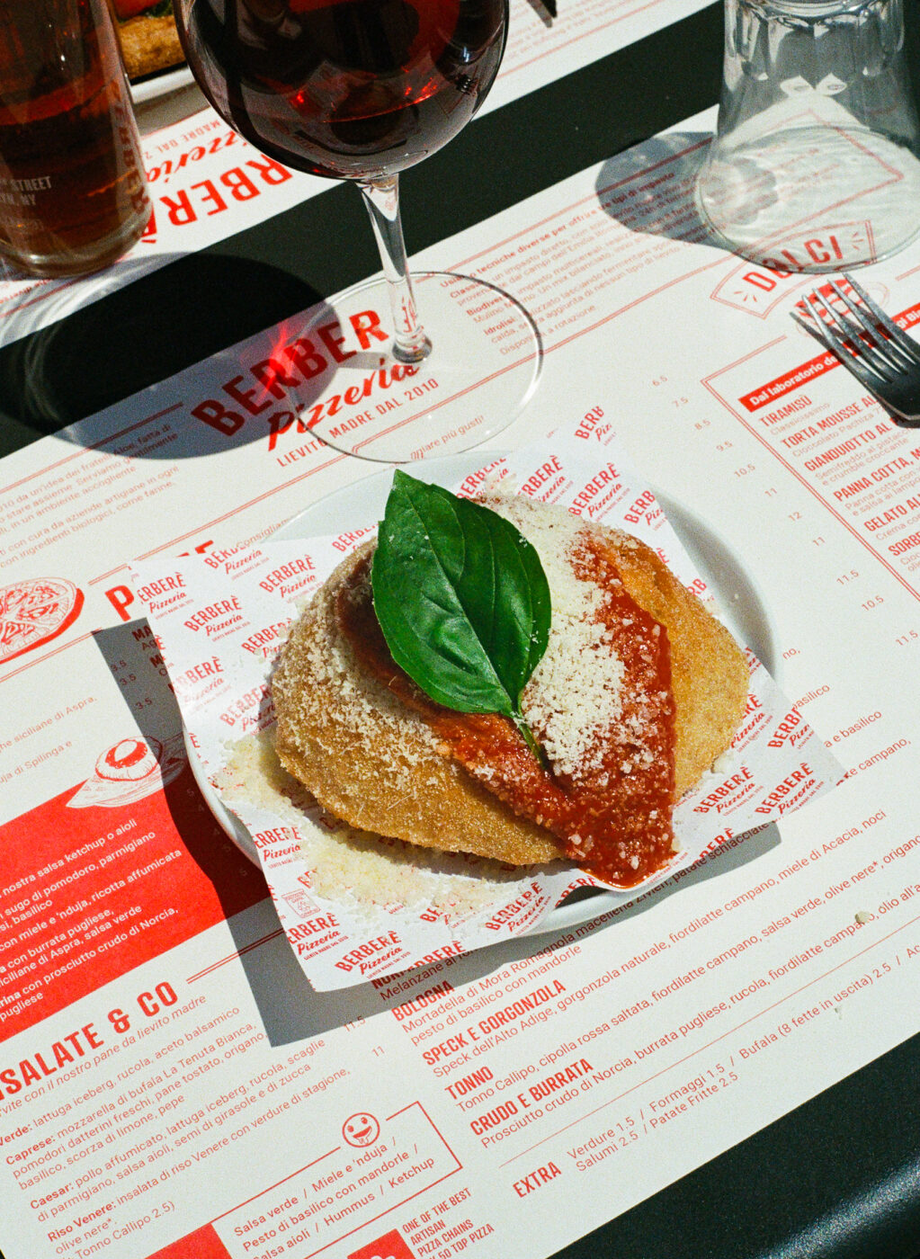 [Nessun titolo dell'immagine] - Un piatto impanato e fritto condito con salsa di pomodoro, formaggio grattugiato e basilico fresco viene servito su un piatto bianco accanto a un bicchiere di vino rosso, su un tavolo di un ristorante con un sottopiatto con il menu stampato.