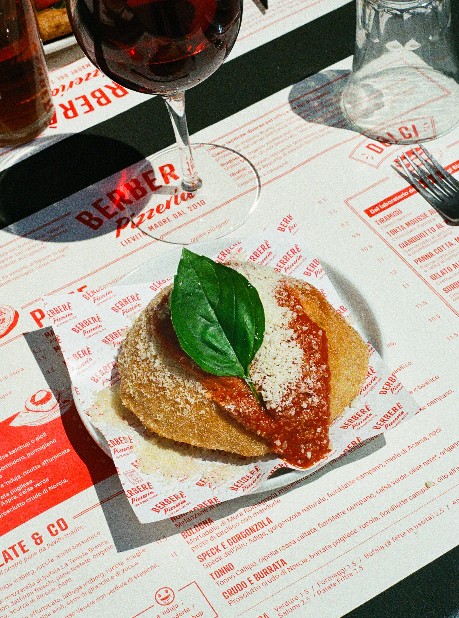 A breaded and fried Italian pizza topped with tomato sauce, grated cheese, and fresh basil sits on a paper tray next to a glass of red wine on a restaurant placemat. If you provide a different image title, let me know and Ill tailor it!.