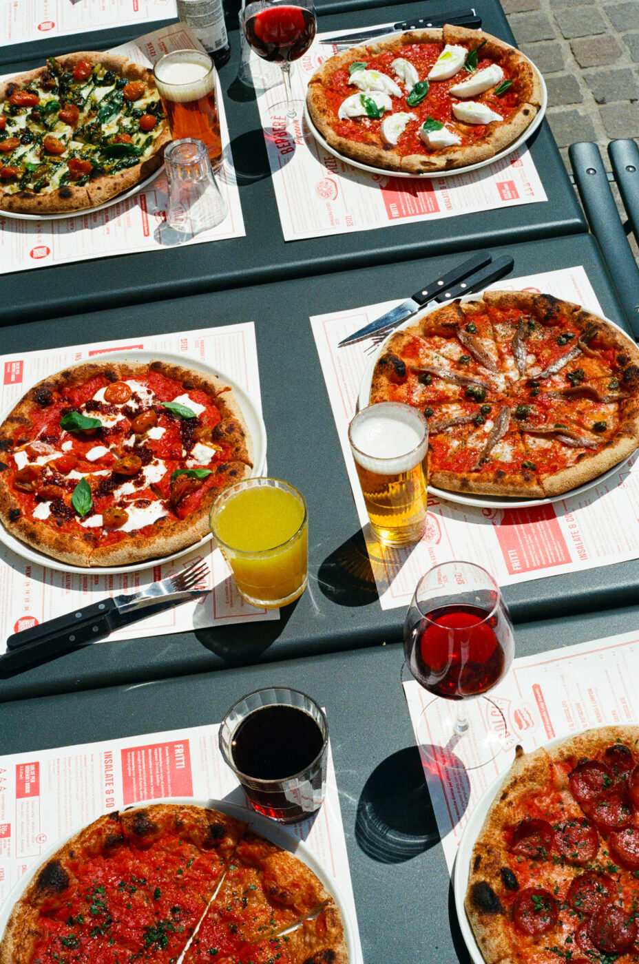 A table outside set with five pizzas, glasses of red and white wine, beer, and orange juice, with utensils and menus arranged underneath each plate. If you specify an actual image title (for example: Italian Dinner Party), let me know and I can adjust it accordingly.