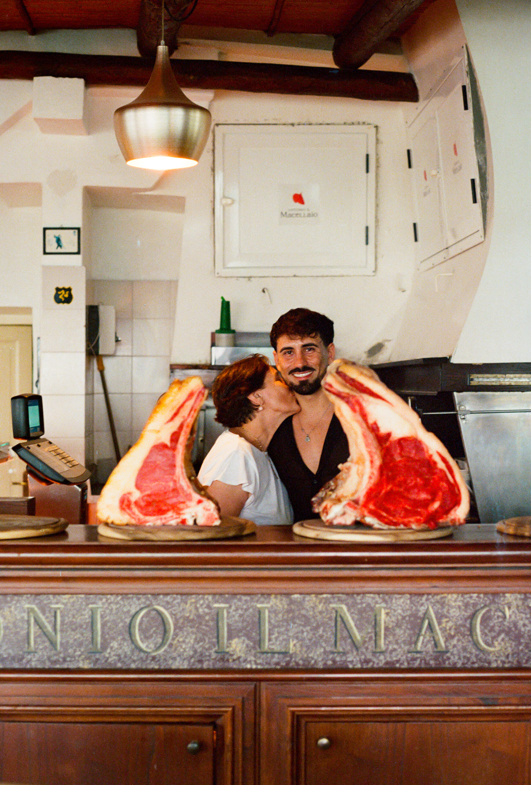 Antonio il Macellaio - A mother kisses her smiling son on the cheek behind a butcher counter with two large raw beef rib cuts displayed in front of them. The shop has white tiled walls and warm lighting.