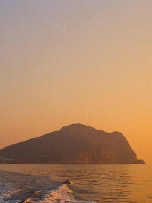 A small, rocky island silhouetted against a golden-orange sky at sunset - A small, rocky island appears in silhouette beneath a glowing orange sky at sunset, viewed from a boat thats leaving a gentle wake across the calm sea.