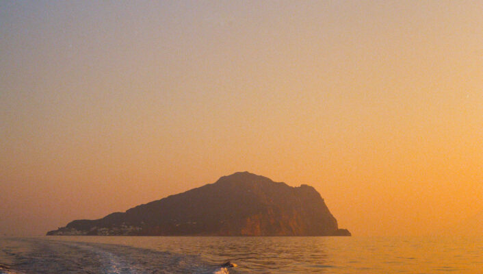 A small, rocky island silhouetted against a golden-orange sky at sunset - A small, rocky island appears in silhouette beneath a glowing orange sky at sunset, viewed from a boat thats leaving a gentle wake across the calm sea.