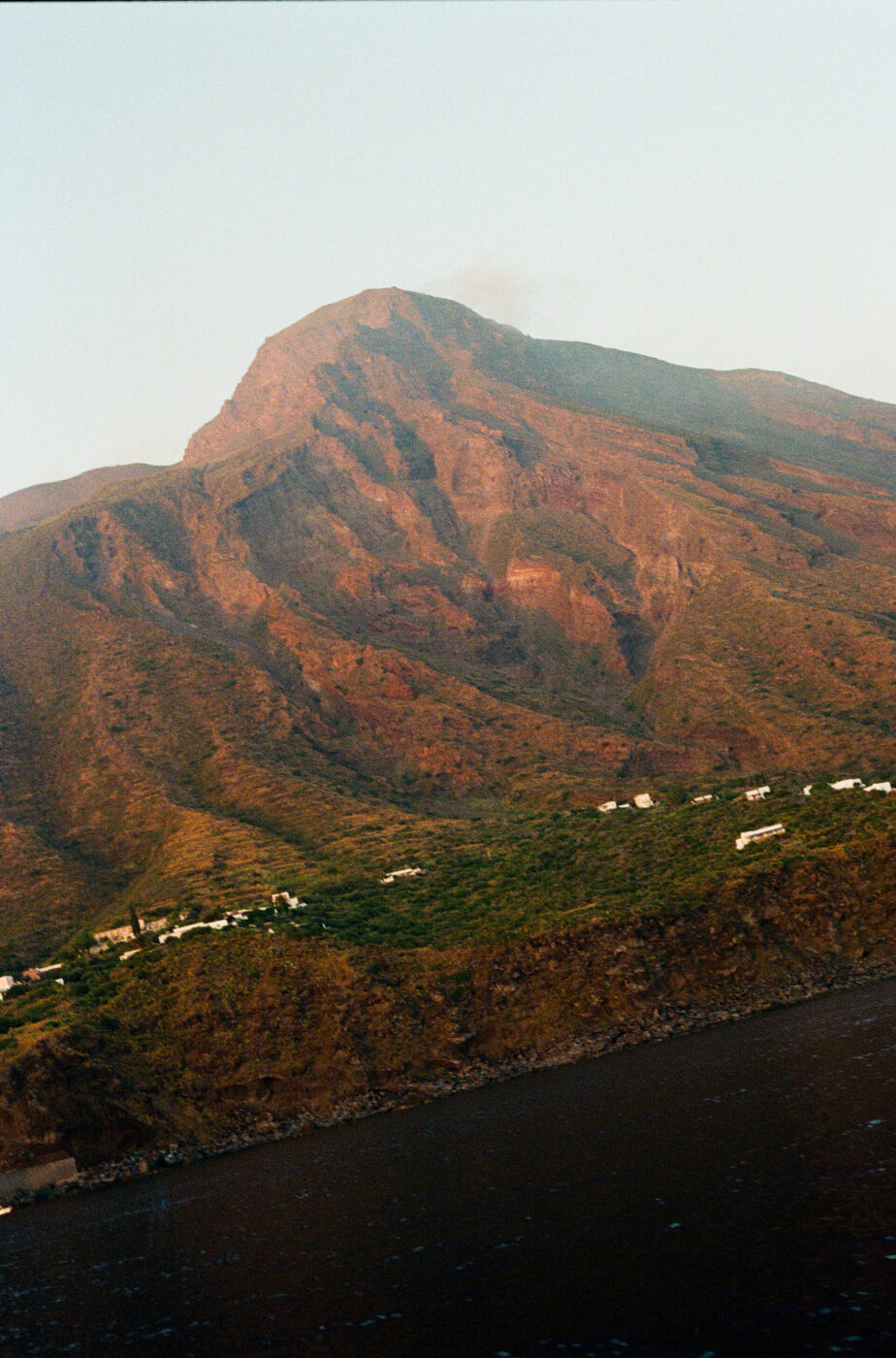 A rugged, reddish-brown volcanic mountain rises above green slopes dotted with small white buildings, overlooking a dark body of water under a hazy sky.