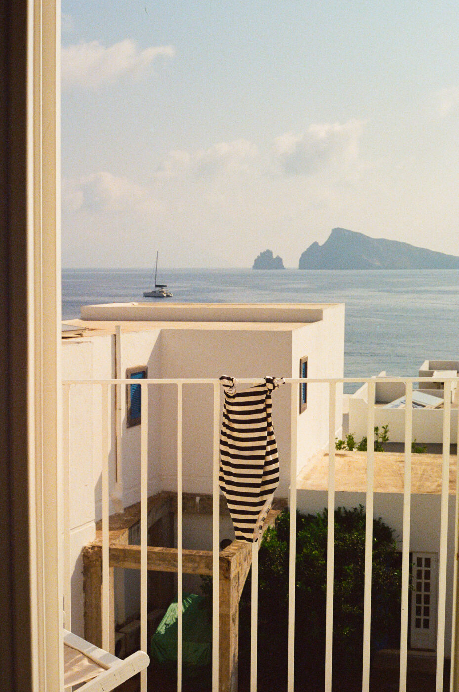 A black and white striped dress hangs over a white balcony railing, overlooking white buildings, calm sea, a distant sailboat, and a rocky island under a clear sky.