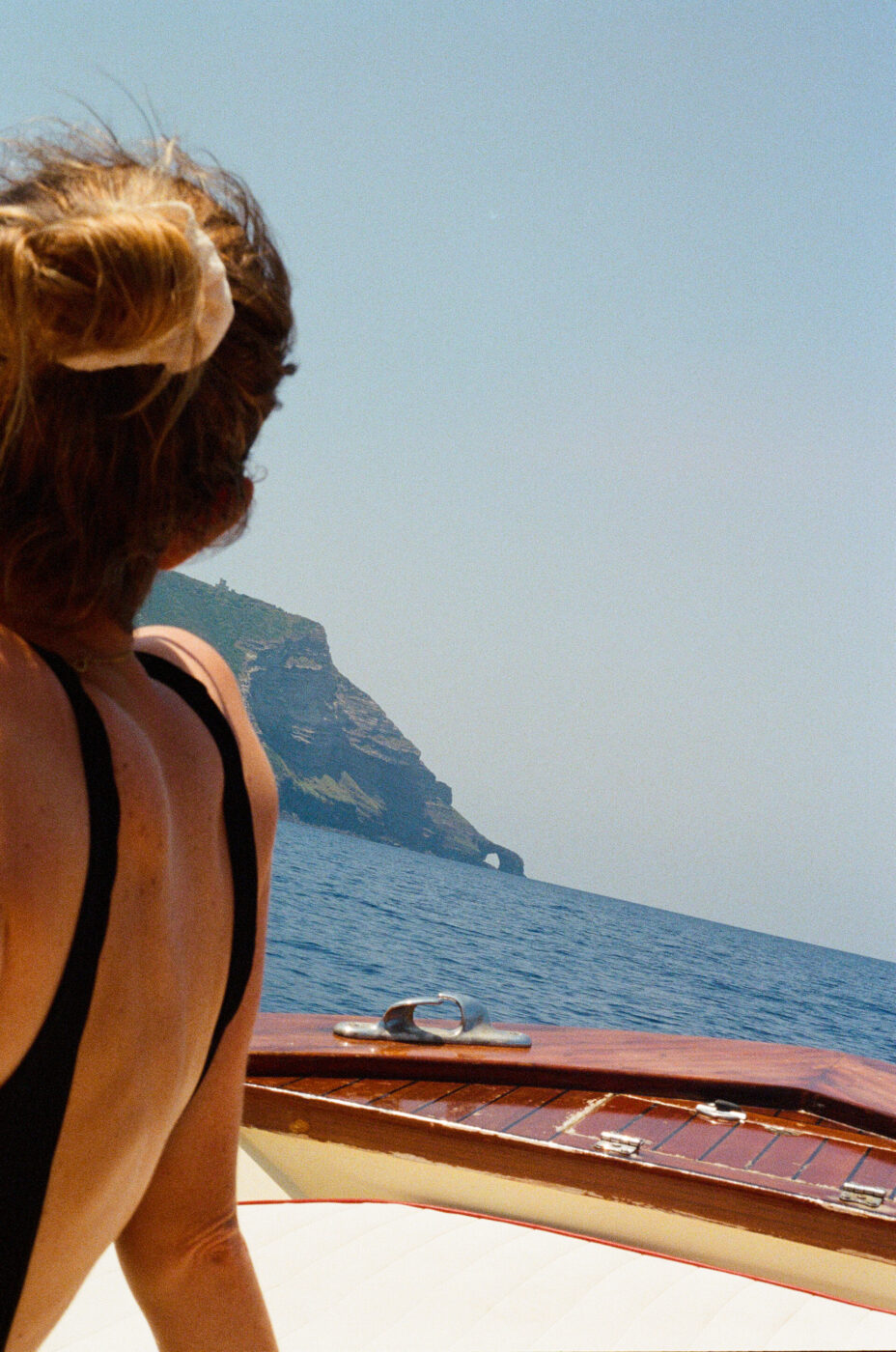 A woman with a scrunchie in her hair and wearing a swimsuit sits on a boat, looking towards the sea and a rocky coastline with a small natural arch in the distance under a clear sky.