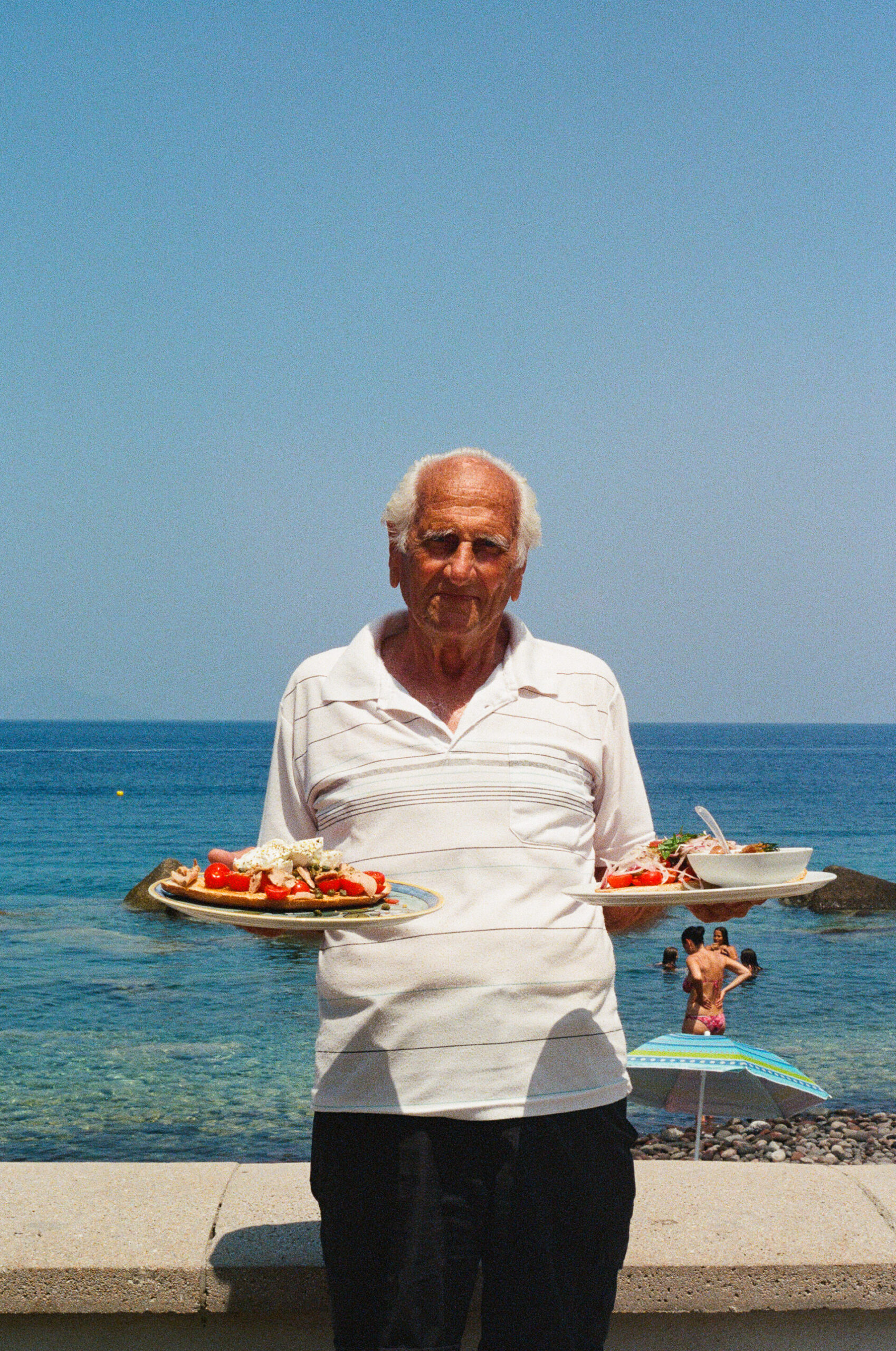 An elderly man in a white polo shirt holds two plates of food while standing by the seaside. The blue ocean and sky are in the background, with a sunbather and colorful umbrella visible behind him. If an image title was provided (let’s say Seaside Lunch), then according to your instruction, the alt text would be formatted as: Seaside Lunch - An elderly man in a white polo shirt holds two plates of food while standing by the seaside. The blue ocean and sky are in the background, with a sunbather and colorful umbrella visible behind him.