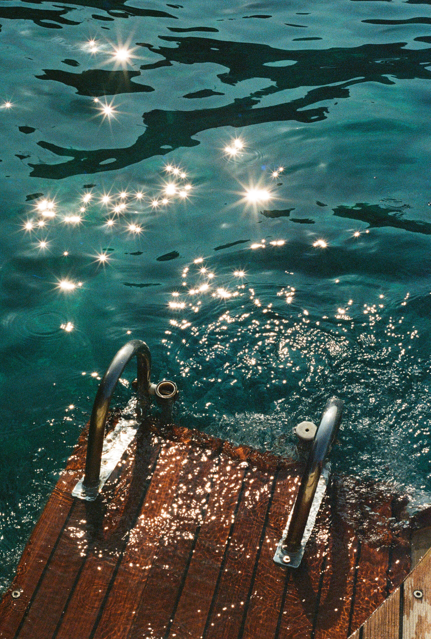 A wooden dock with metal handrails leads into rippling blue-green water, reflecting bright sunlight that creates sparkling star-like patterns on the surface.