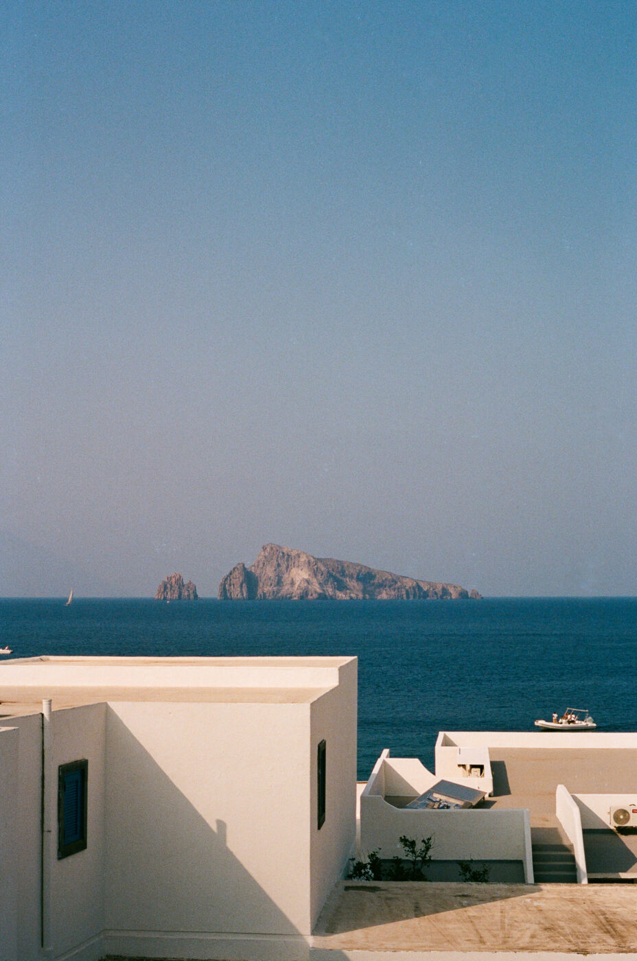 Panarea - White Mediterranean buildings with blue accents are in the foreground, overlooking a calm blue sea. In the distance, a rocky island rises from the water under a clear sky, with a few boats scattered nearby.