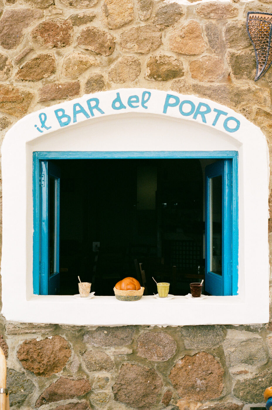 Il BAR del PORTO - A stone wall with a blue-framed open window labeled il BAR del PORTO. Three cups with spoons and a bread basket sit on the windowsill, suggesting a café or bar setting.
