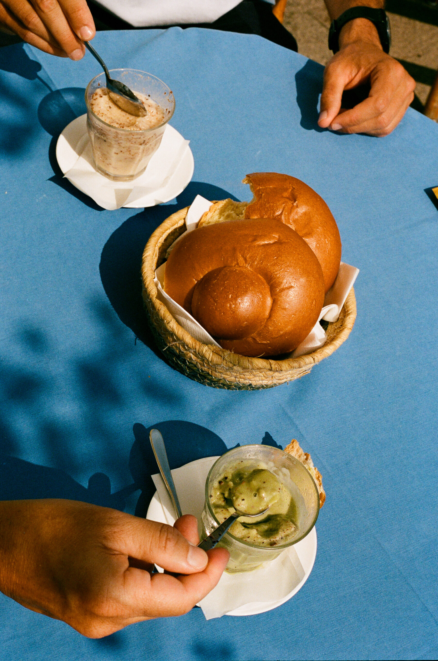 Two people sit at a blue table with a basket of brioche bread and two glass cups of dessert, one with green granita and one with a creamy dessert. Each person holds a spoon, ready to eat.