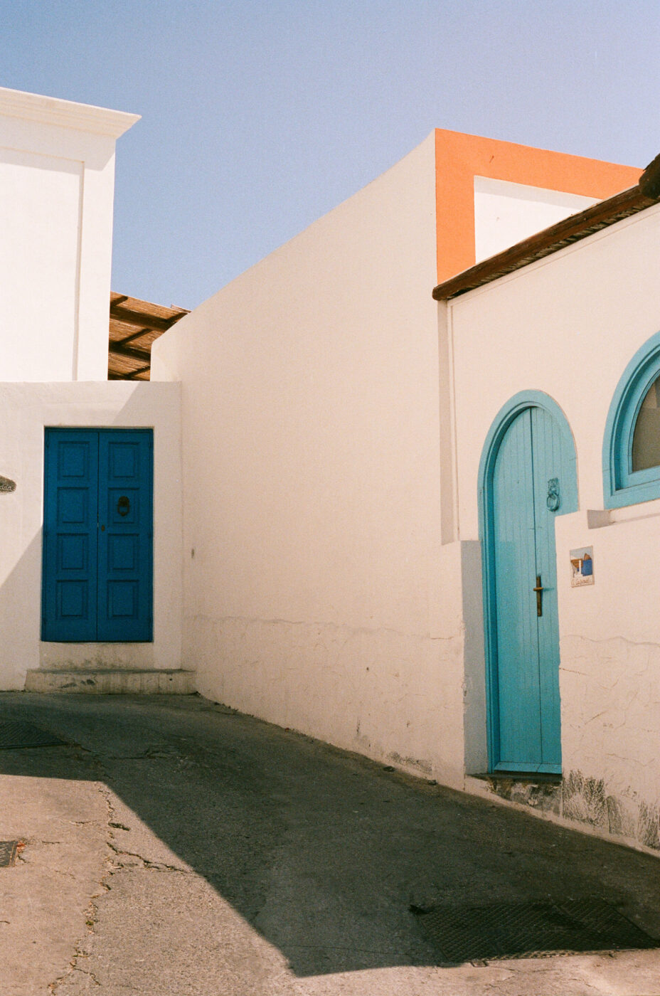 A narrow street with white buildings featuring bright blue doors and window frames, under a clear sky - A narrow street lined with white buildings that have bright blue doors and window frames, bathed in sunlight on a clear day, creating a peaceful Mediterranean atmosphere.