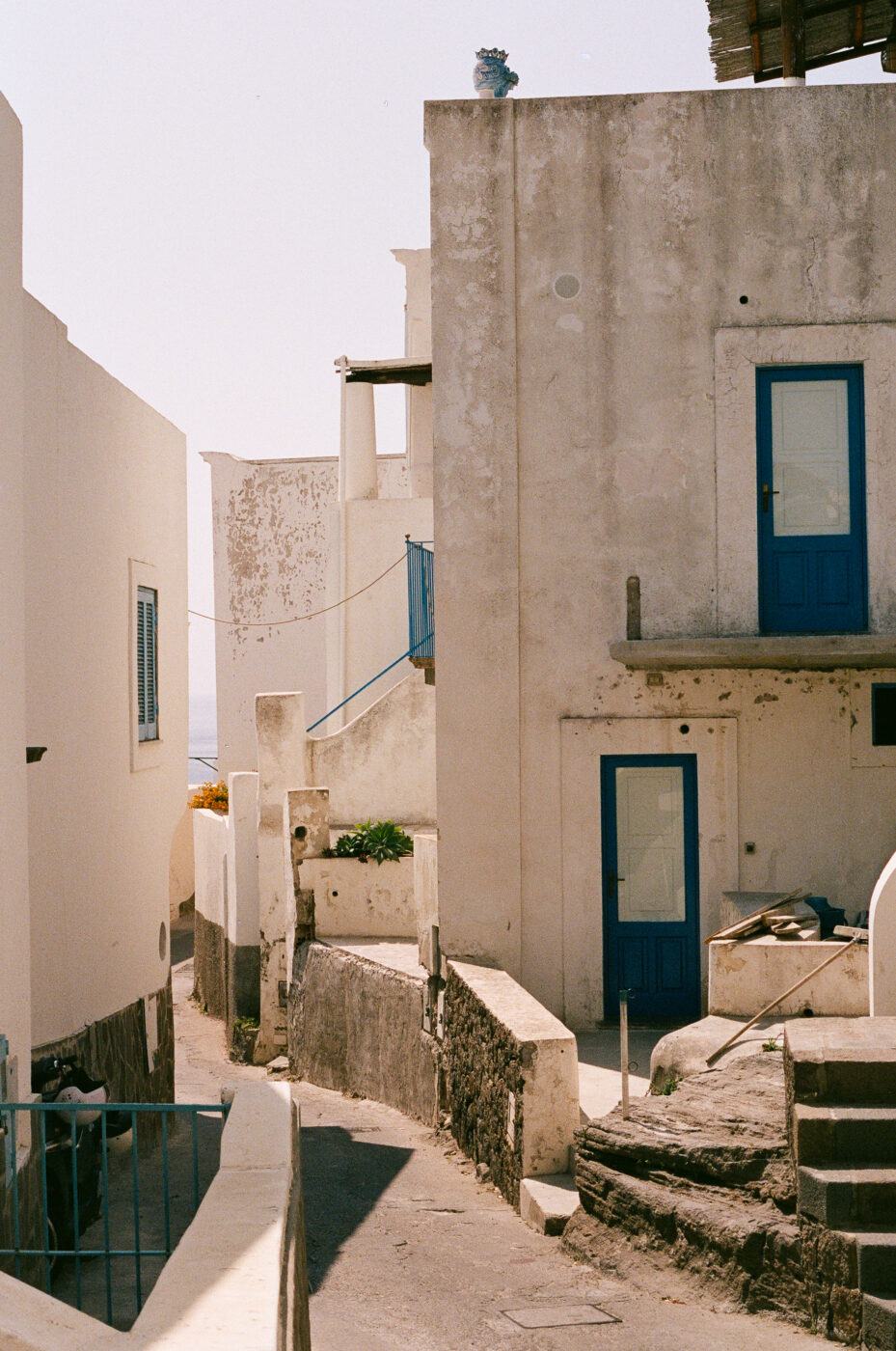 Vicolo stretto in un villaggio mediterraneo - Un vicolo stretto si snoda tra edifici imbiancati con porte e finestre blu in un soleggiato villaggio mediterraneo. Le ombre proiettate sui muri aggiungono contrasto alla scena.