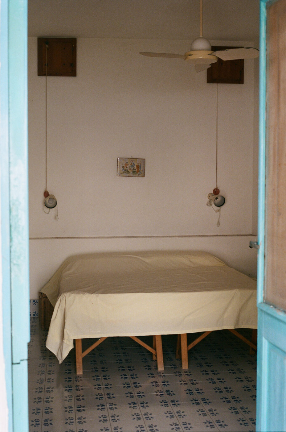 A cozy bedroom with a bed covered in a light yellow blanket, patterned tile floor, hanging pendant lights, a ceiling fan, and a small framed artwork on the white wall seen through an open doorway.