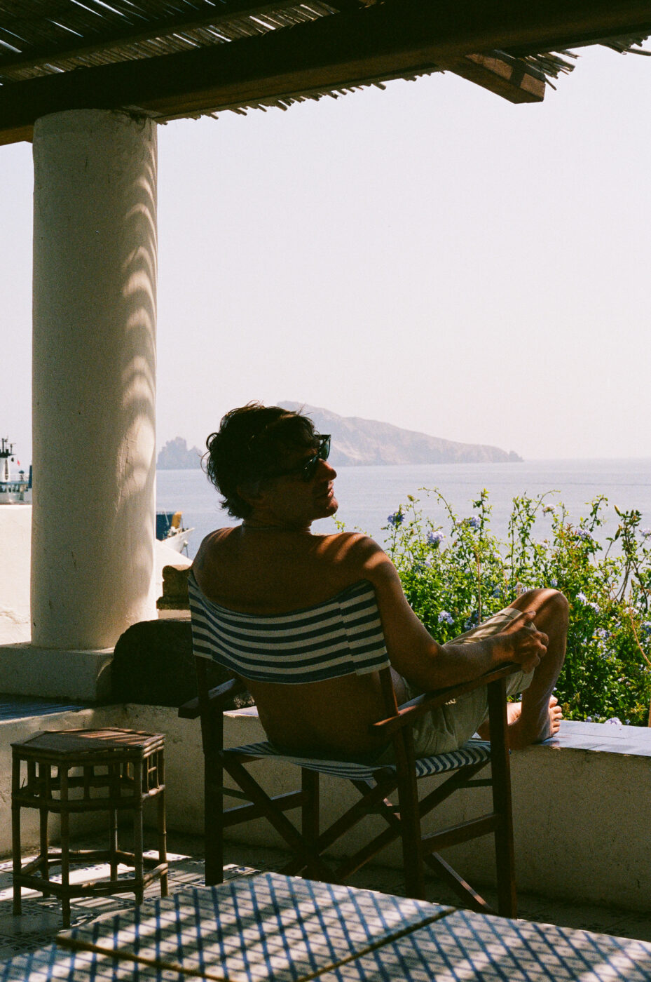 Seaside Relaxation - A person in sunglasses and a striped outfit sits on a shaded terrace, relaxing in a chair and looking out at the sea and distant island on a sunny day. If no image title is provided, simply use the descriptive part: A person in sunglasses and a striped outfit sits on a shaded terrace, relaxing in a chair and looking out at the sea and distant island on a sunny day.