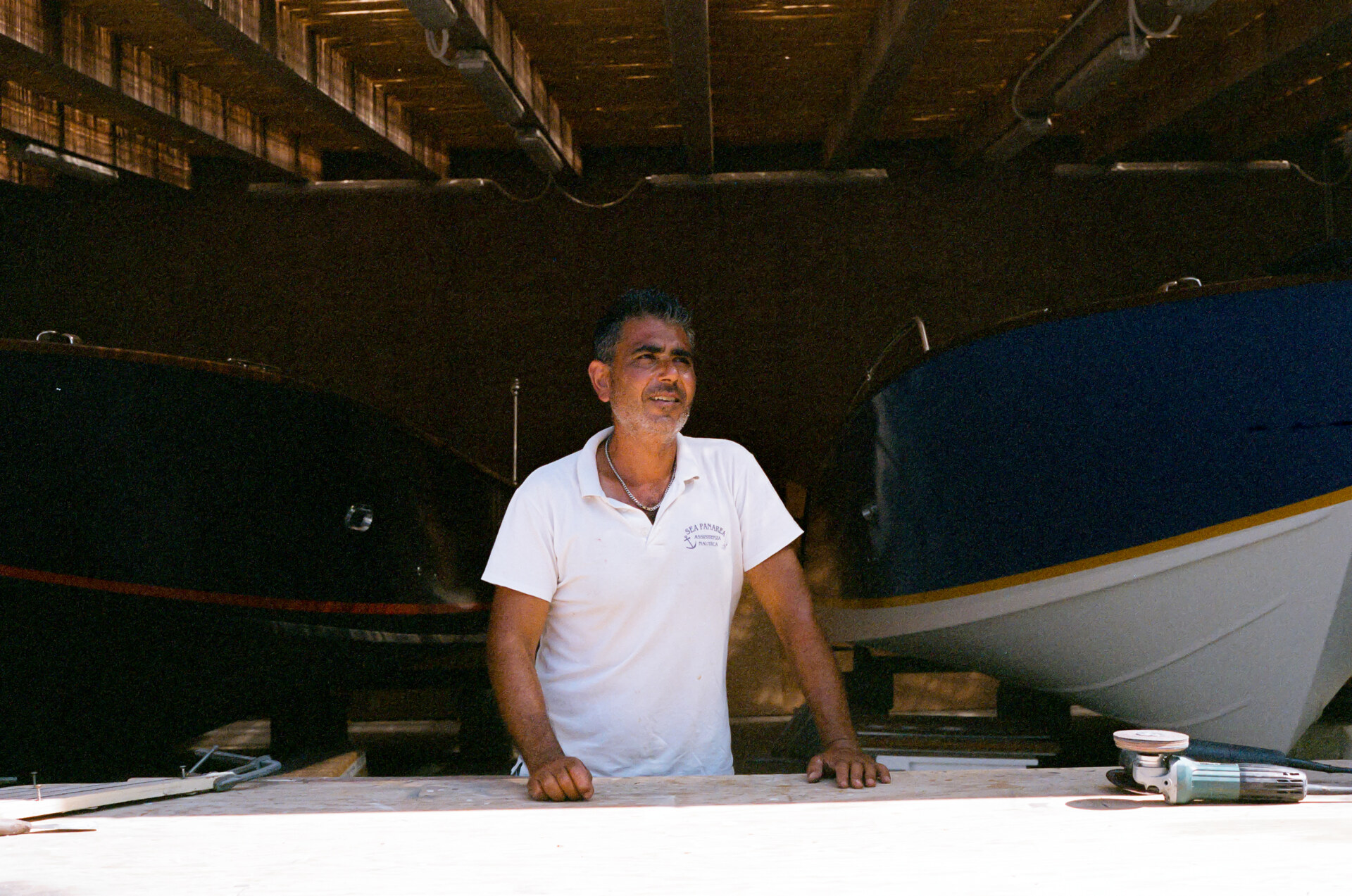 Boat Builder at Work - A man in a white polo shirt stands at a wooden workbench inside a boat shed, with two boats behind him and sunlight streaming through the roof. If youd like alt texts for other images or have a specific title in mind, let me know!.