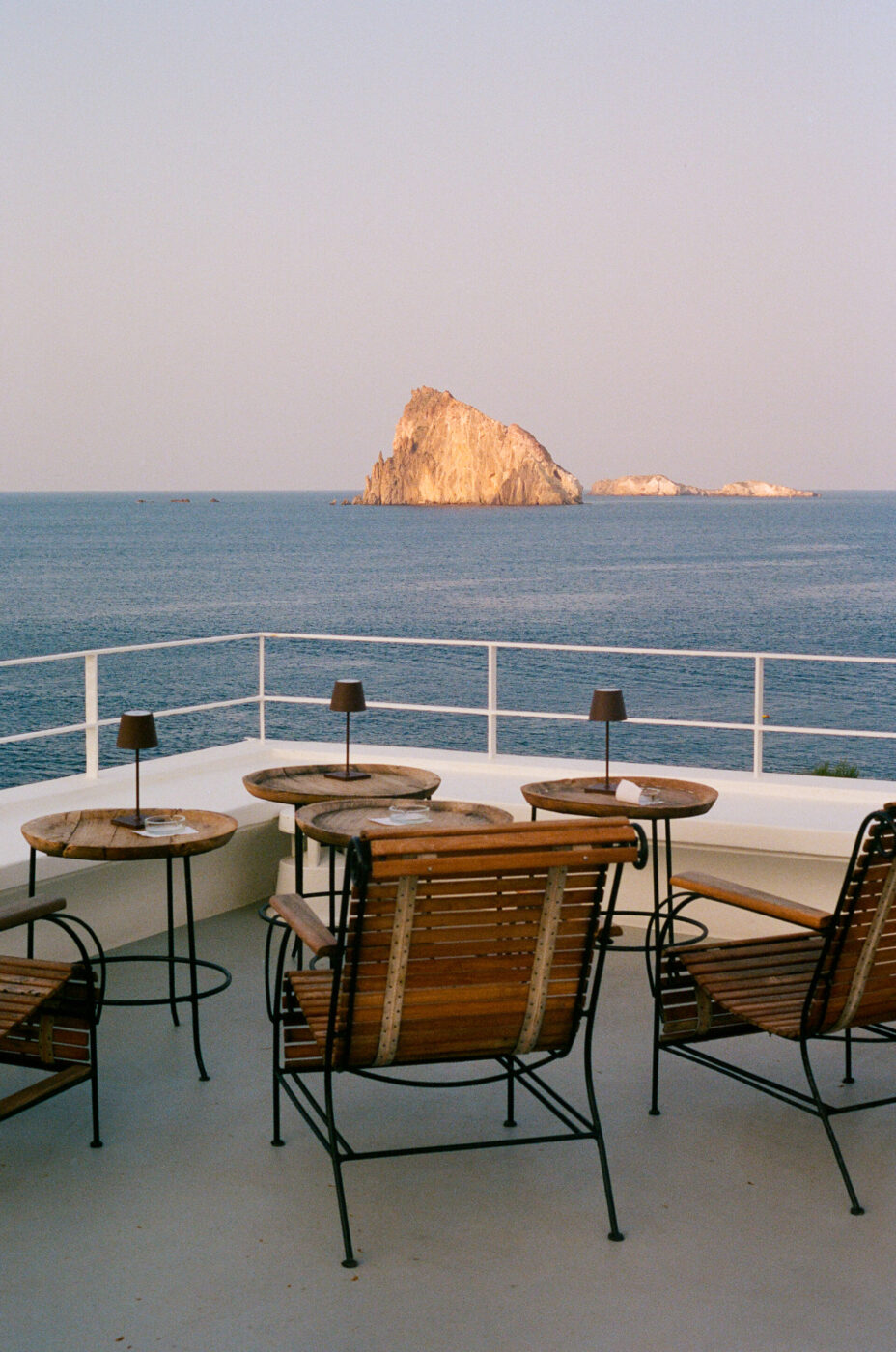Wooden chairs and tables on seaside terrace - Wooden chairs and tables are arranged on a terrace overlooking the ocean, with a rocky island visible in the distance under a clear sky.