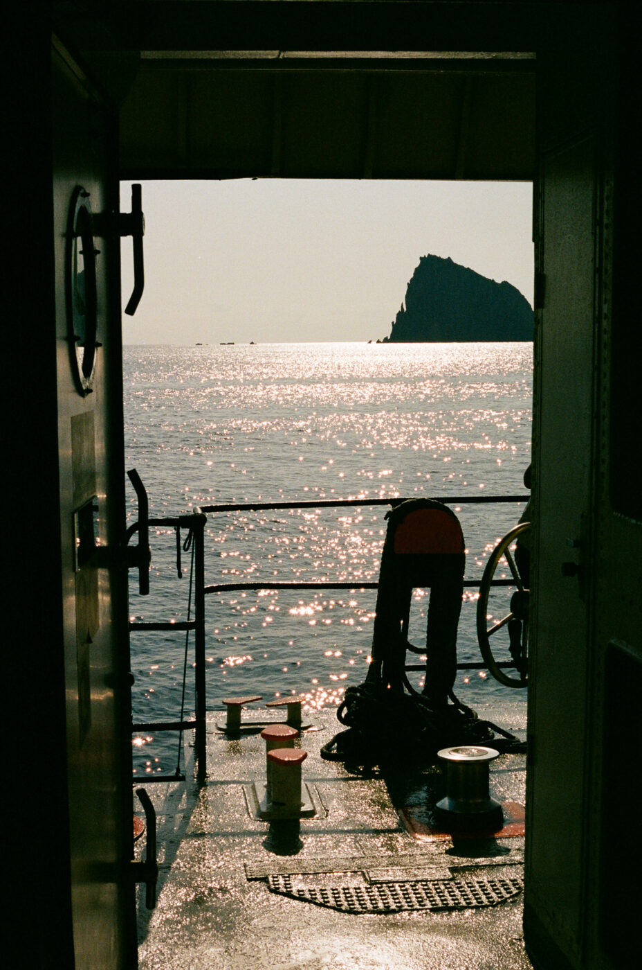 Vista dall'interno di una nave che si affaccia su un mare scintillante e illuminato dal sole, con un'isola o uno sperone roccioso visibile in lontananza; le attrezzature della nave e le ringhiere si stagliano in primo piano.