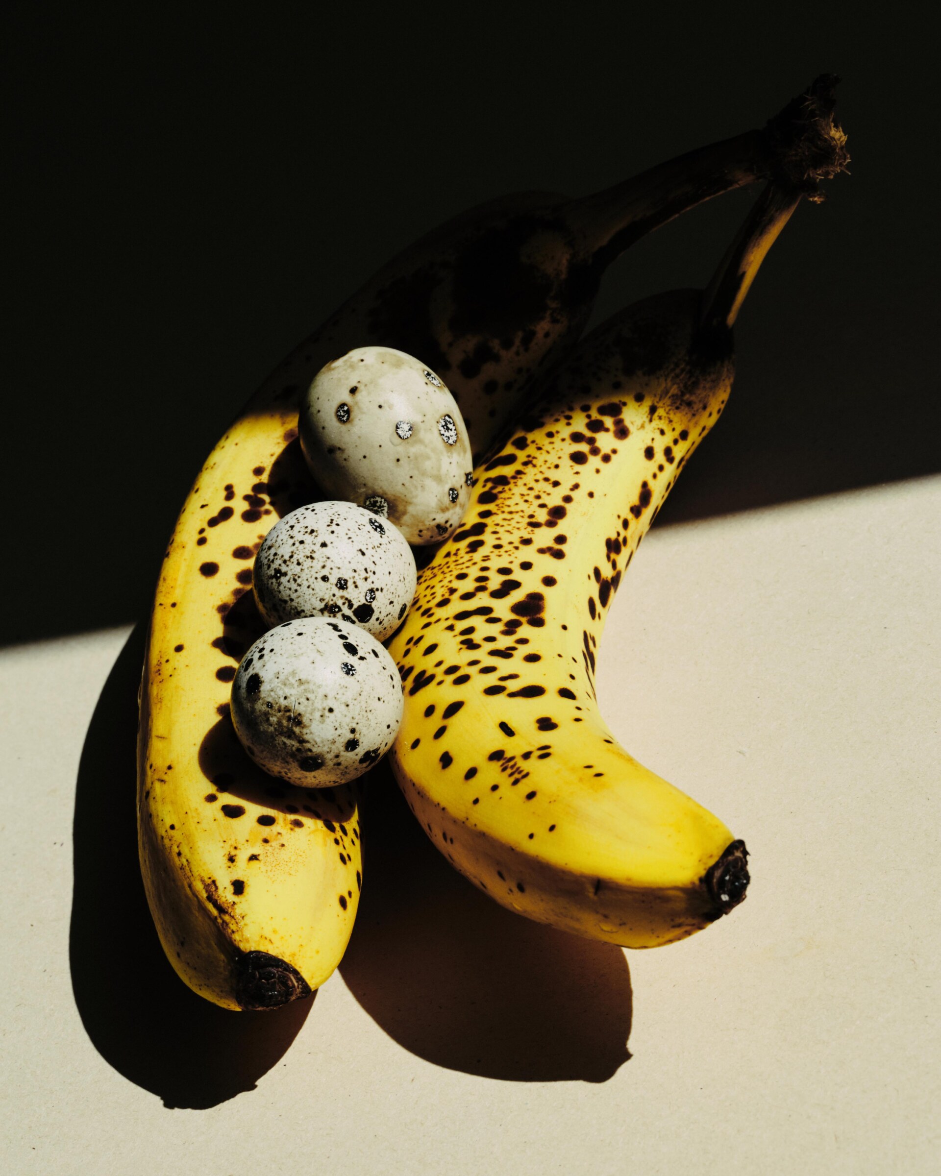 Due banane mature con macchie marroni e tre uova di quaglia punteggiate riposano su una superficie chiara, parzialmente illuminata dalla luce del sole, con le ombre che aggiungono contrasto.
