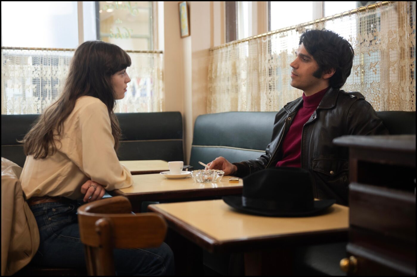 Poiché non è stato fornito un titolo per l'immagine, il testo alternativo appropriato sarebbe: Una donna e un uomo sono seduti uno di fronte all'altro a un tavolino di un bar, impegnati a conversare. La donna indossa una camicetta color crema, l'uomo una giacca scura e sul tavolo ci sono un cappello nero e delle tazze da caffè.