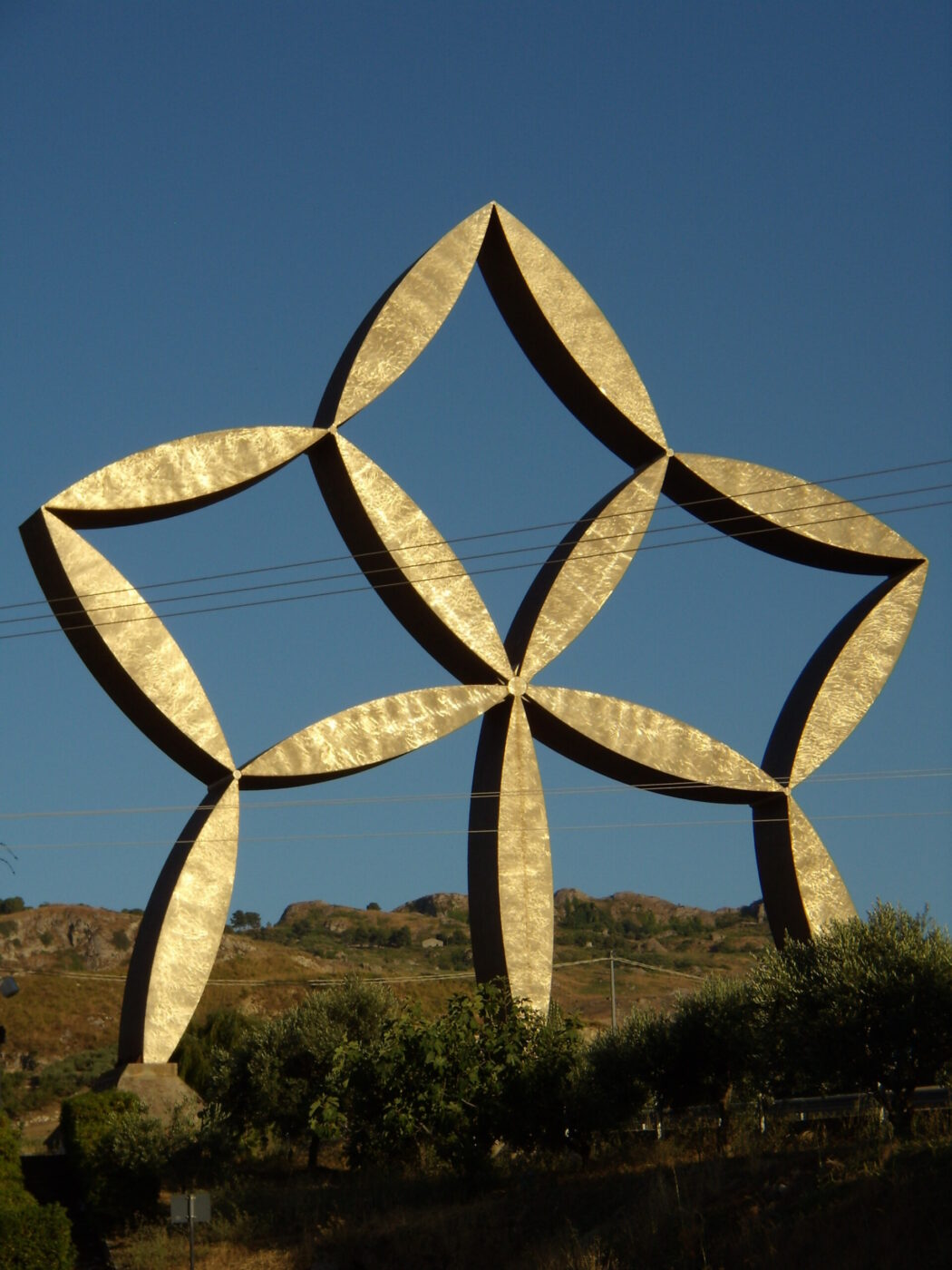 Una grande scultura dorata a forma di stella, composta da travi curve che si intersecano, si erge all'aperto in un cielo azzurro e limpido, con colline e alberi sullo sfondo.