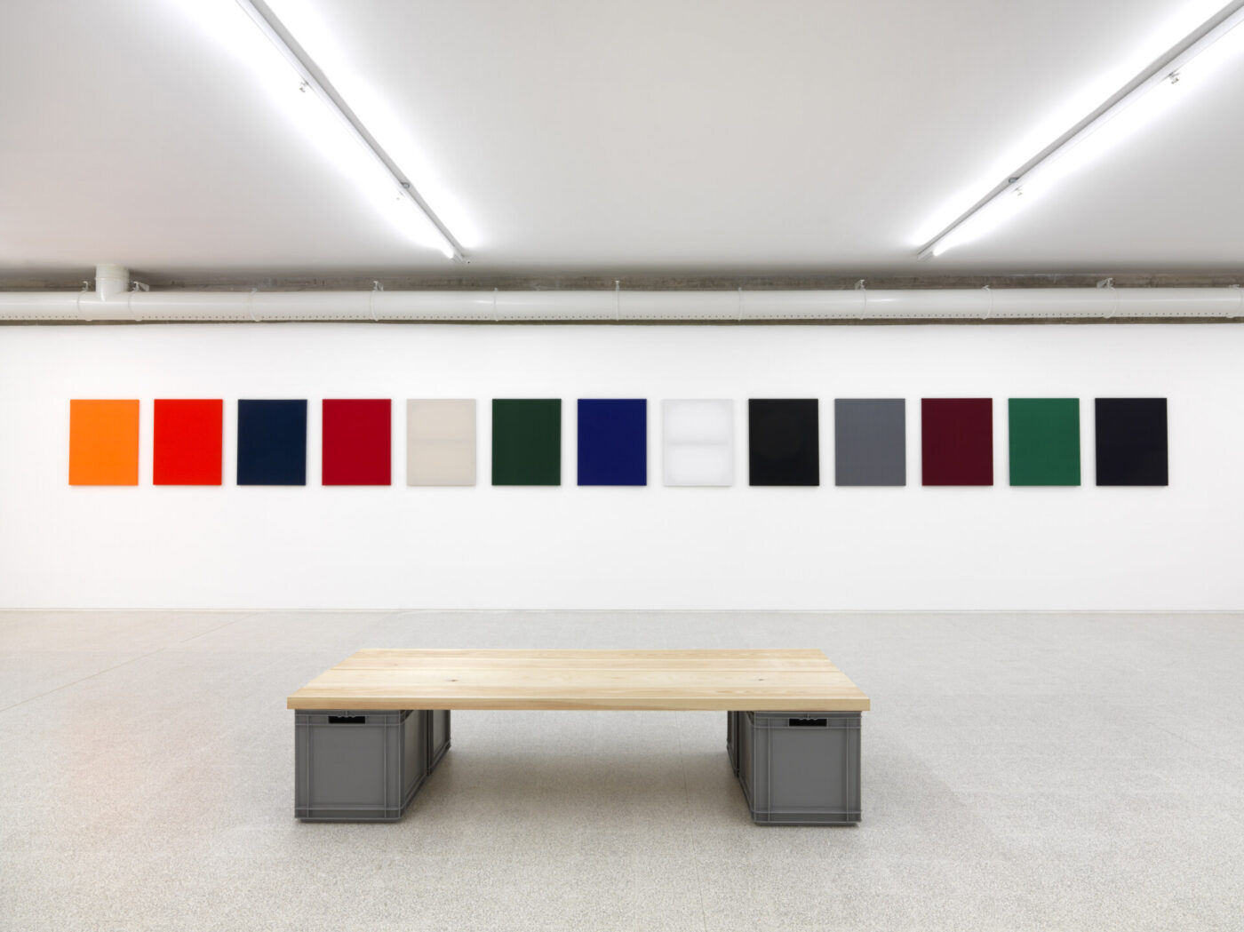 A minimalist gallery with a wooden bench in the center, facing a wall displaying twelve rectangular abstract artworks of varying colors and patterns, all evenly spaced in a row.