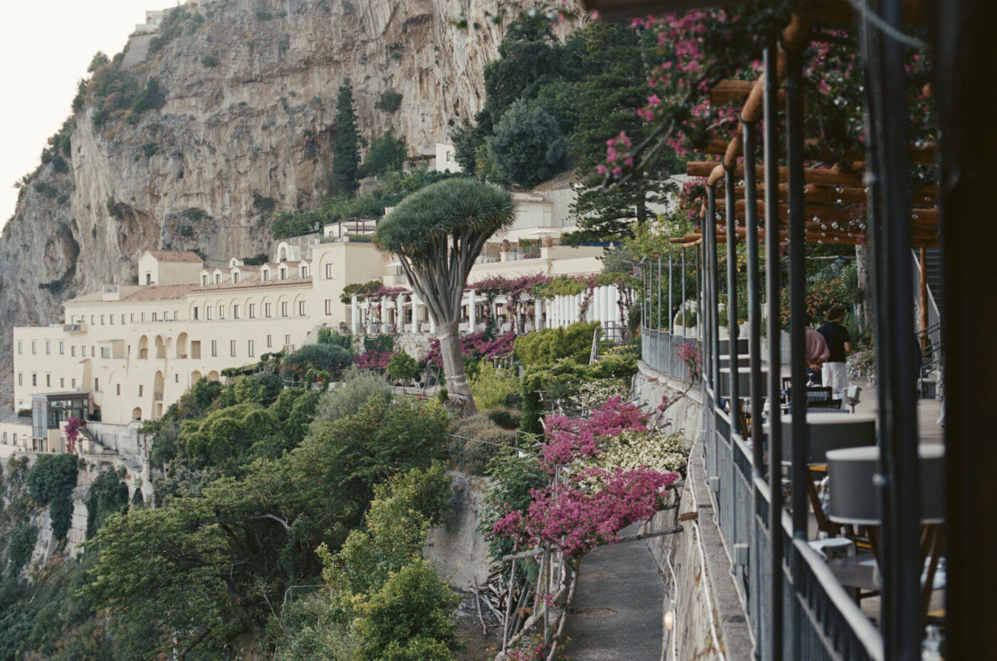 Certo! Ecco il testo alt con il titolo dell'immagine posto all'inizio, come da istruzioni: Una terrazza collinare con tavoli e sedie si affaccia su un paesaggio lussureggiante, fiori rosa in fiore ed edifici color crema costruiti su ripide scogliere. Alberi e vegetazione circondano la scenografica area montuosa. Tuttavia, nel prompt non è stato fornito il titolo dell'immagine. Se è presente un titolo dell'immagine (ad esempio: "Vista sulla Costiera Amalfitana"), il testo alt dovrebbe essere formattato come segue: Vista sulla Costiera Amalfitana - Una terrazza in collina con tavoli e sedie si affaccia su un paesaggio lussureggiante, fiori rosa in fiore ed edifici color crema costruiti su ripide scogliere. Alberi e verde circondano la scenografica area montuosa. Se mi dite il titolo specifico dell'immagine che volete utilizzare, posso aggiornarlo di conseguenza!.