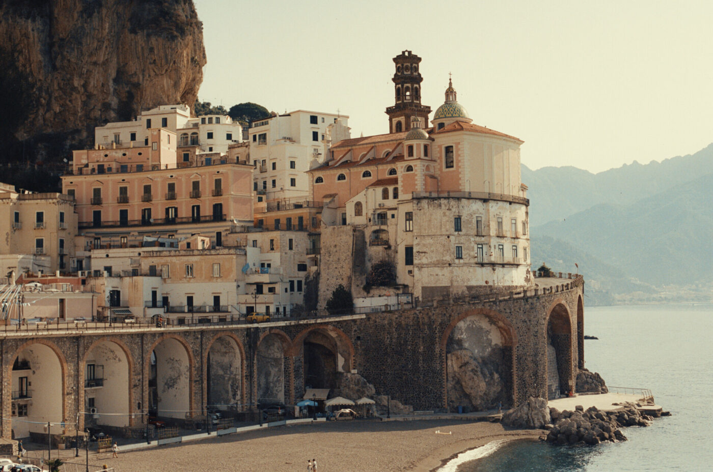 Edifici colorati in riva al mare con sostegni ad arco si trovano su una scogliera sopra una spiaggia sabbiosa e un mare azzurro e calmo, con le montagne sullo sfondo sotto un cielo limpido.