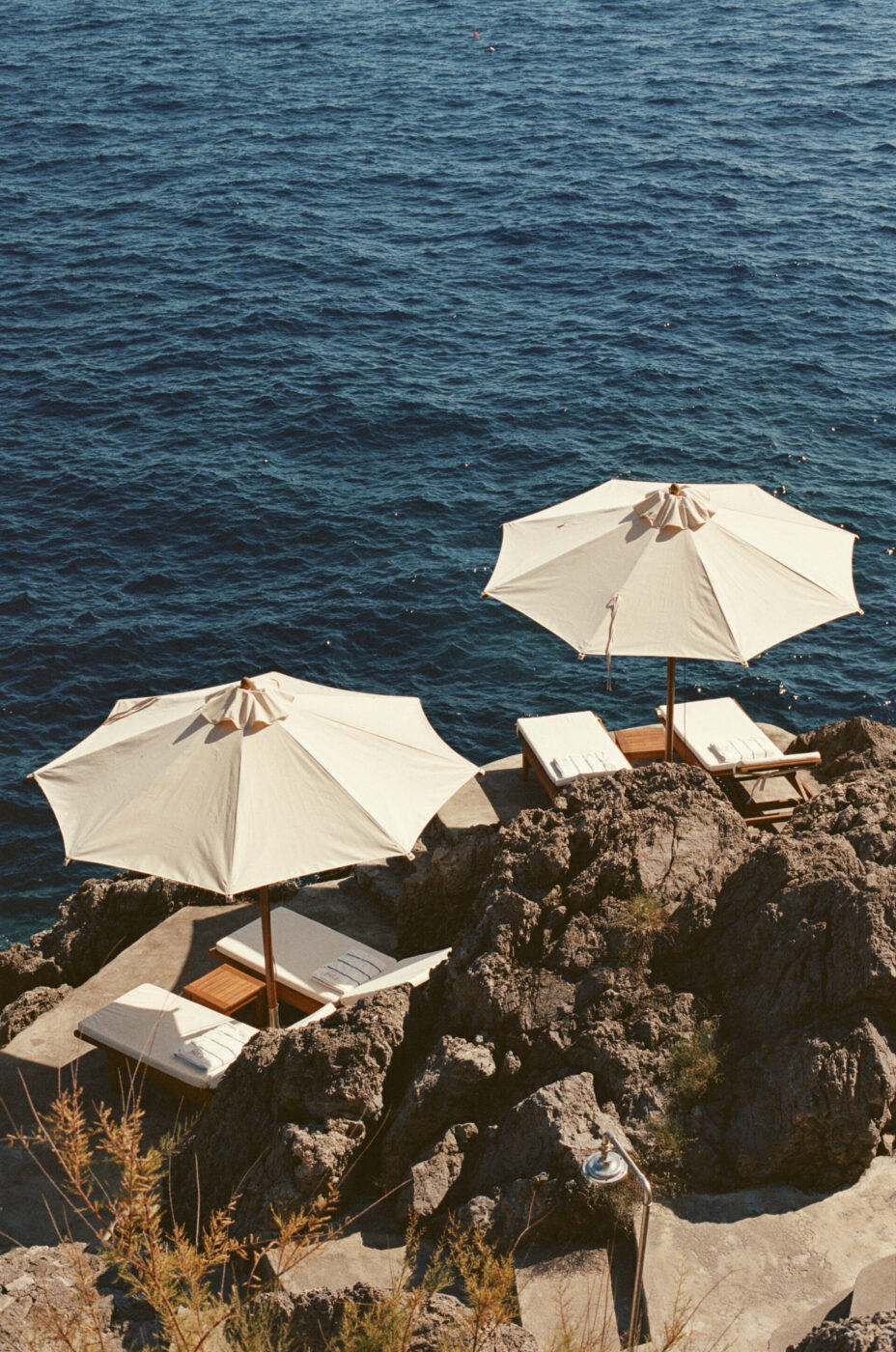 [Titolo dell'immagine] - Due ombrelloni bianchi e lettini sono sistemati su scogliere rocciose che si affacciano su un mare azzurro brillante, creando un tranquillo angolo di relax in riva al mare.