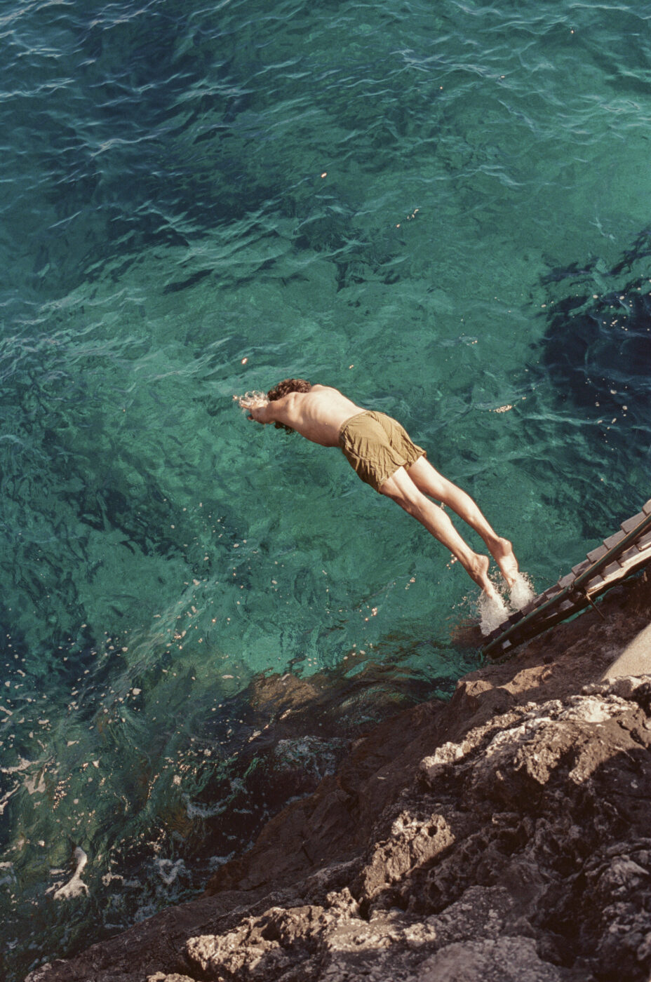 Titolo dell'immagine: Cliff Dive - Una persona in pantaloncini abbronzati si tuffa da una sporgenza rocciosa in un'acqua chiara e turchese, catturata a mezz'aria sopra il mare.