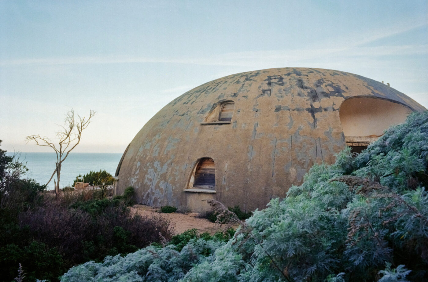 A weathered, dome-shaped building with arched windows stands near a coastline, surrounded by green shrubs and a barren tree, under a pale blue sky.