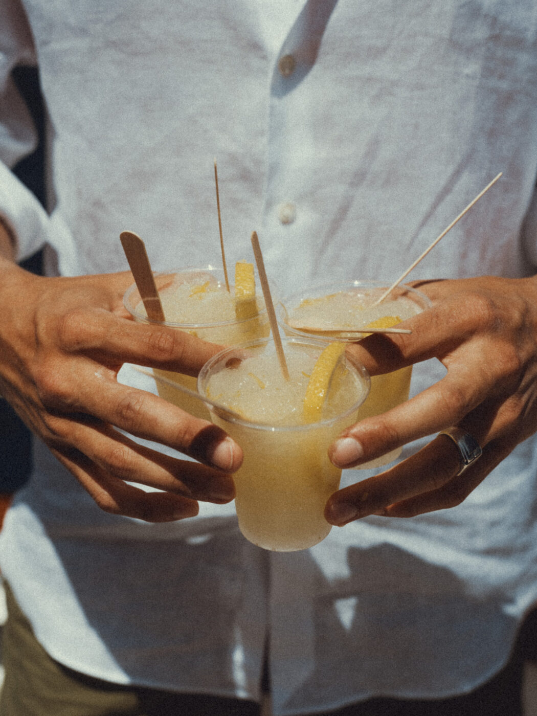 [Una persona in camicia bianca tiene in mano tre tazze di una bevanda gialla ghiacciata guarnita con fette di limone e bastoncini di legno, una tazza in ogni mano e tiene in equilibrio la terza.