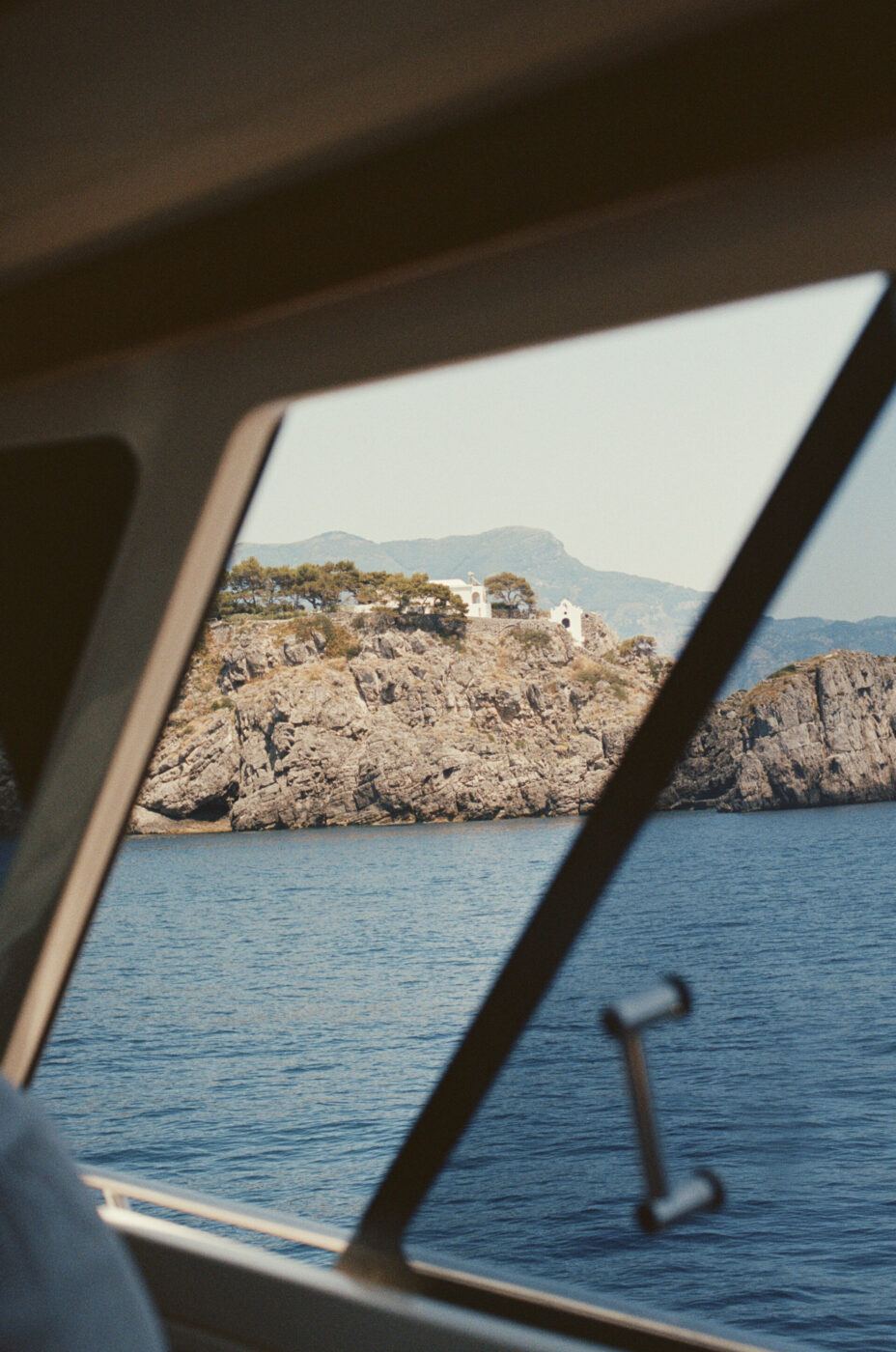 Una costa rocciosa con una casa bianca circondata da alberi - Una costa rocciosa con una casa bianca immersa tra gli alberi, vista attraverso il finestrino di una barca che galleggia su acque calme e blu, con montagne visibili sullo sfondo in lontananza.