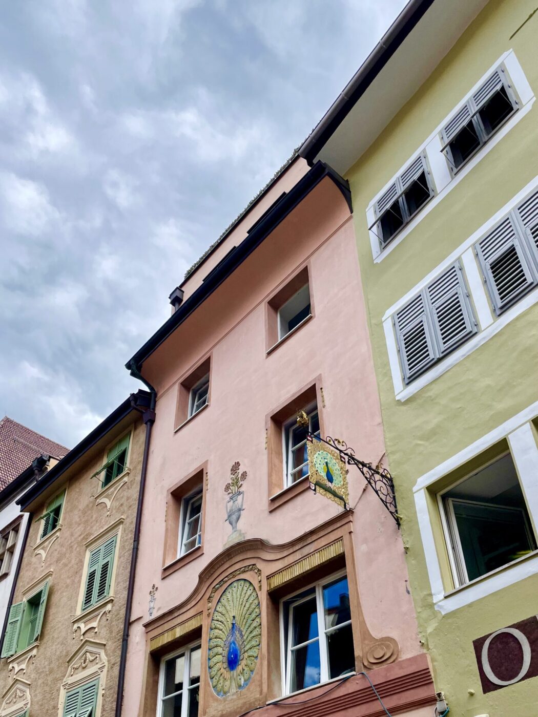 Colorful European Buildings with Peacock Motif - Colorful European buildings with pastel facades and decorative shutters. One building features a painted peacock motif above the entrance and an ornate sign with a peacock, set against a cloudy sky.