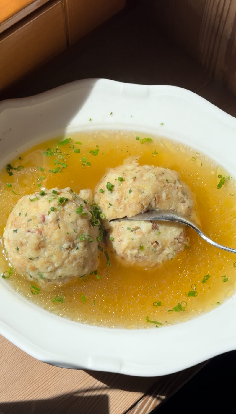 Two Bread Dumplings in Clear Broth - Two bread dumplings served in a clear broth, garnished with chopped fresh chives in a white bowl. A spoon rests next to the dumplings. Sunlight highlights the dish.