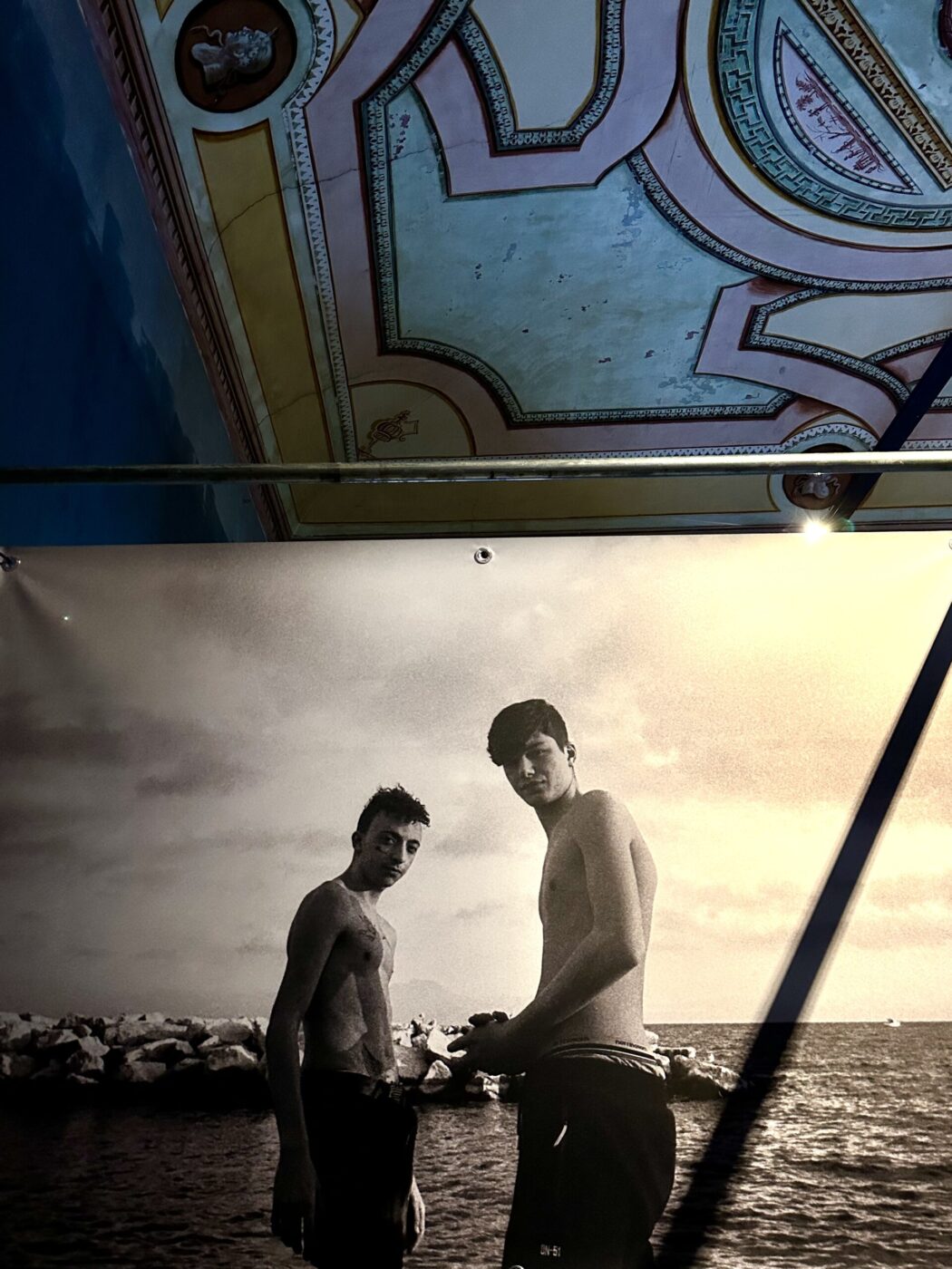 A photo of two young men standing shirtless by the sea - A photo of two young men standing shirtless by the sea is displayed on a wall beneath a decorative, ornate ceiling with classical designs and pastel colors.